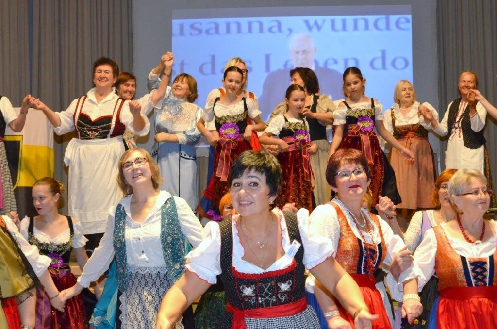 Frauen und Mädchen in Trachten tanzen gemeinsam auf einer Bühne und halten sich an den Händen.