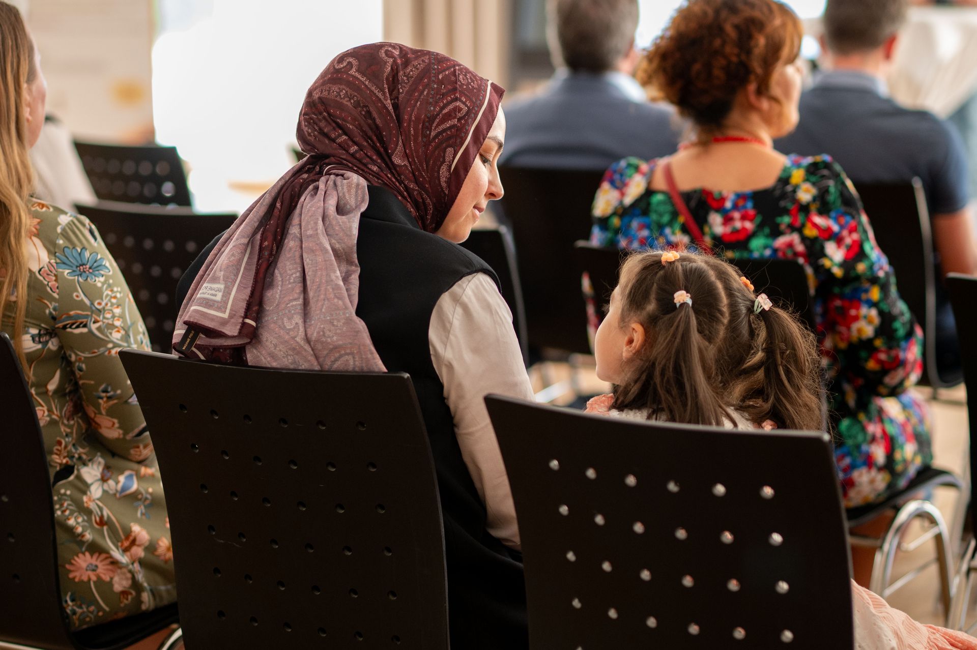 Frau mit Hijab sitzt neben einem kleinen Mädchen bei einer Veranstaltung. Sie schauen sich an.