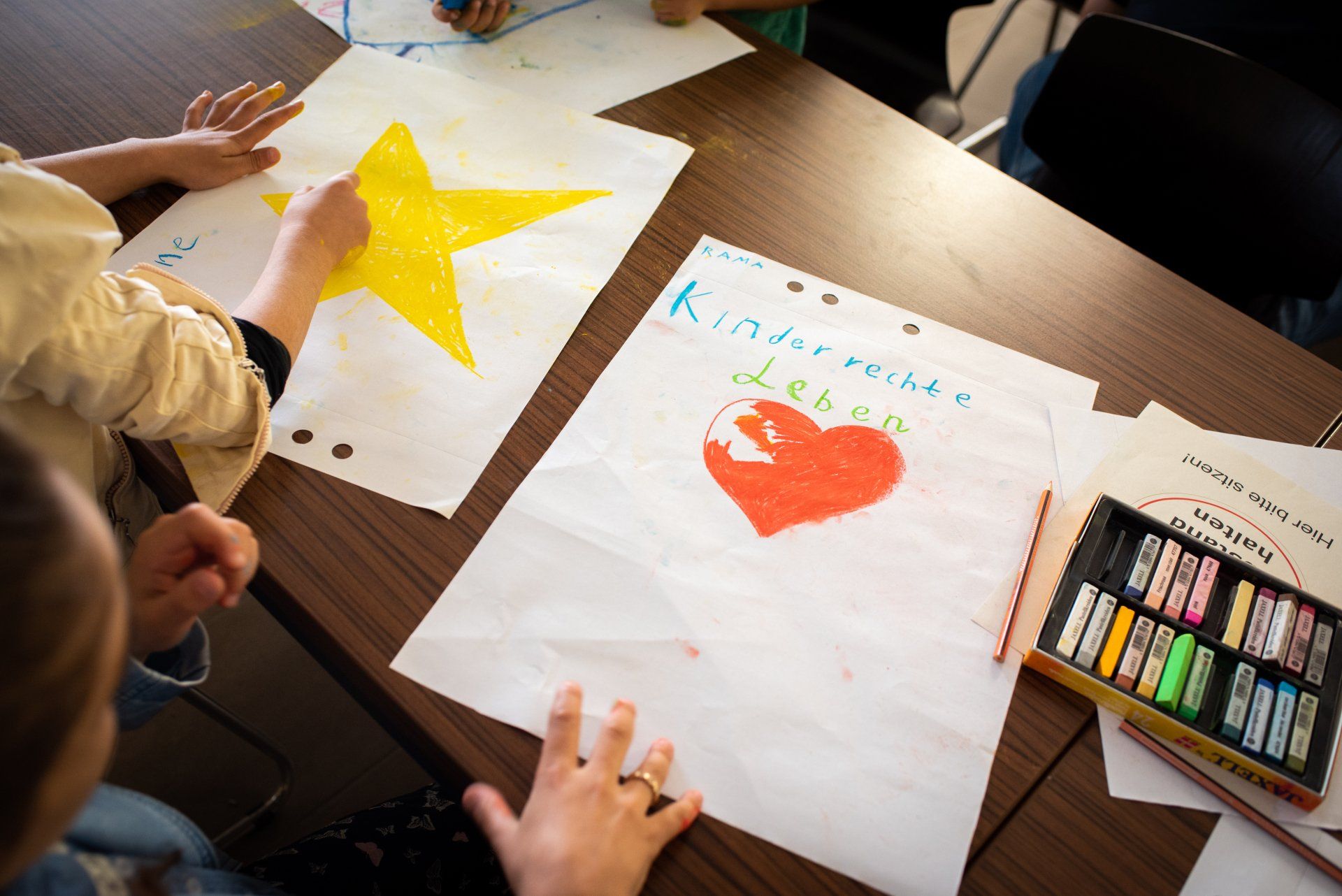 Kinder malen auf zwei großen Blättern. Link sein gelber Stern, rechts ein Herz, darüber