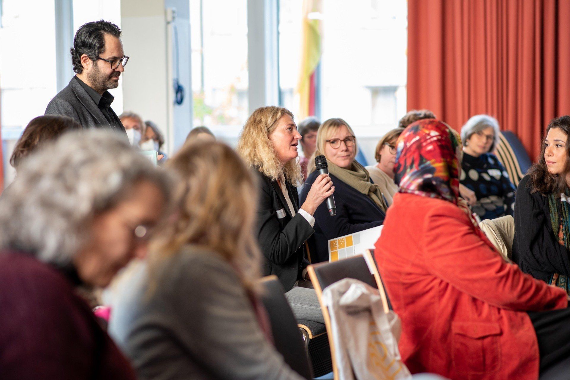 Frau im Plenum, die ins Mikrofon spricht. Um sie herum Eltern und Akteure mit und ohne Einwanderungsgeschichte. Link führt zu Seite der Fachtagung 2022