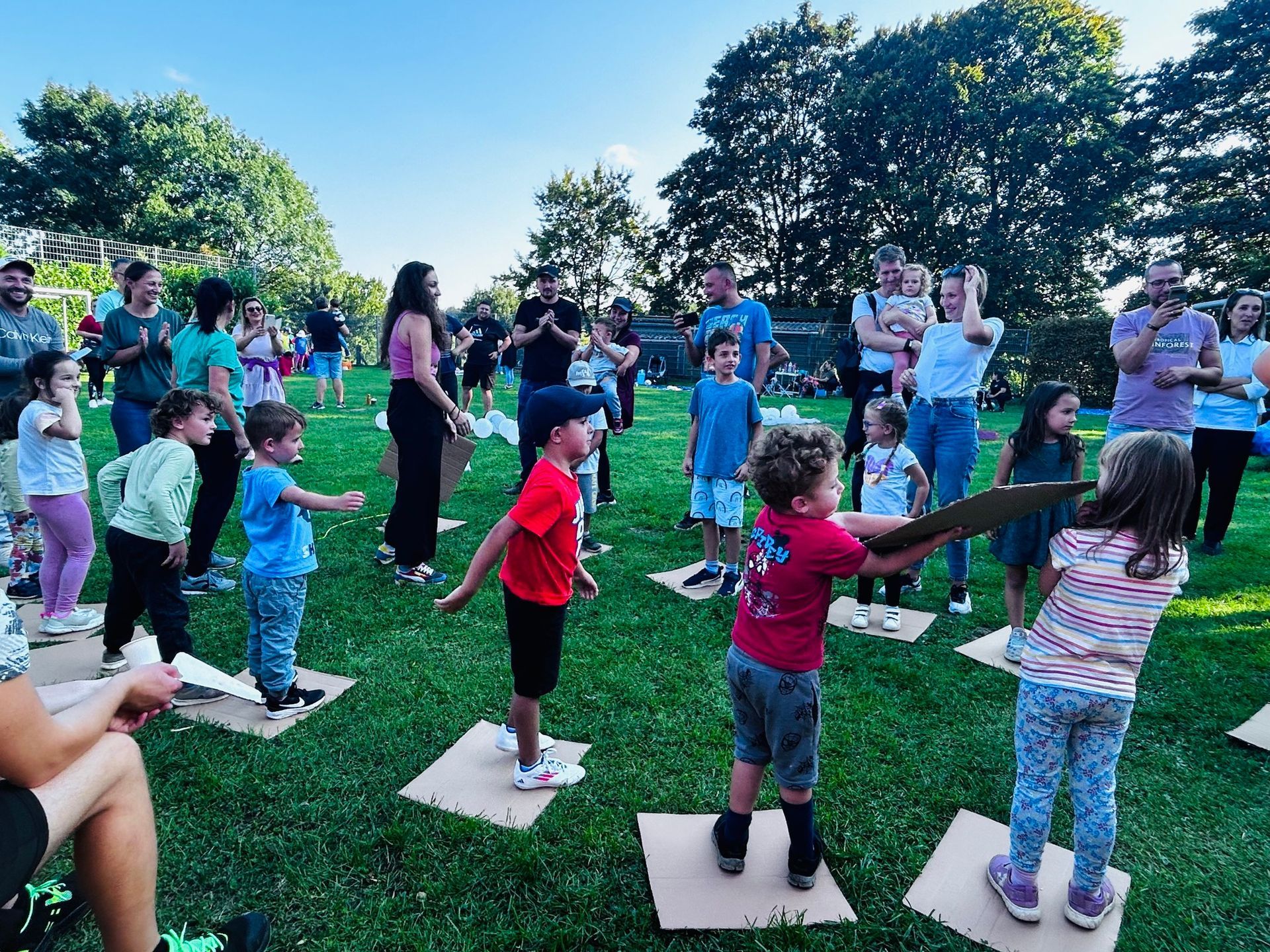 Kinder und Erwachsene spielen auf einer Wiese ein Teamspiel mit Pappunterlagen und Pappstücken.