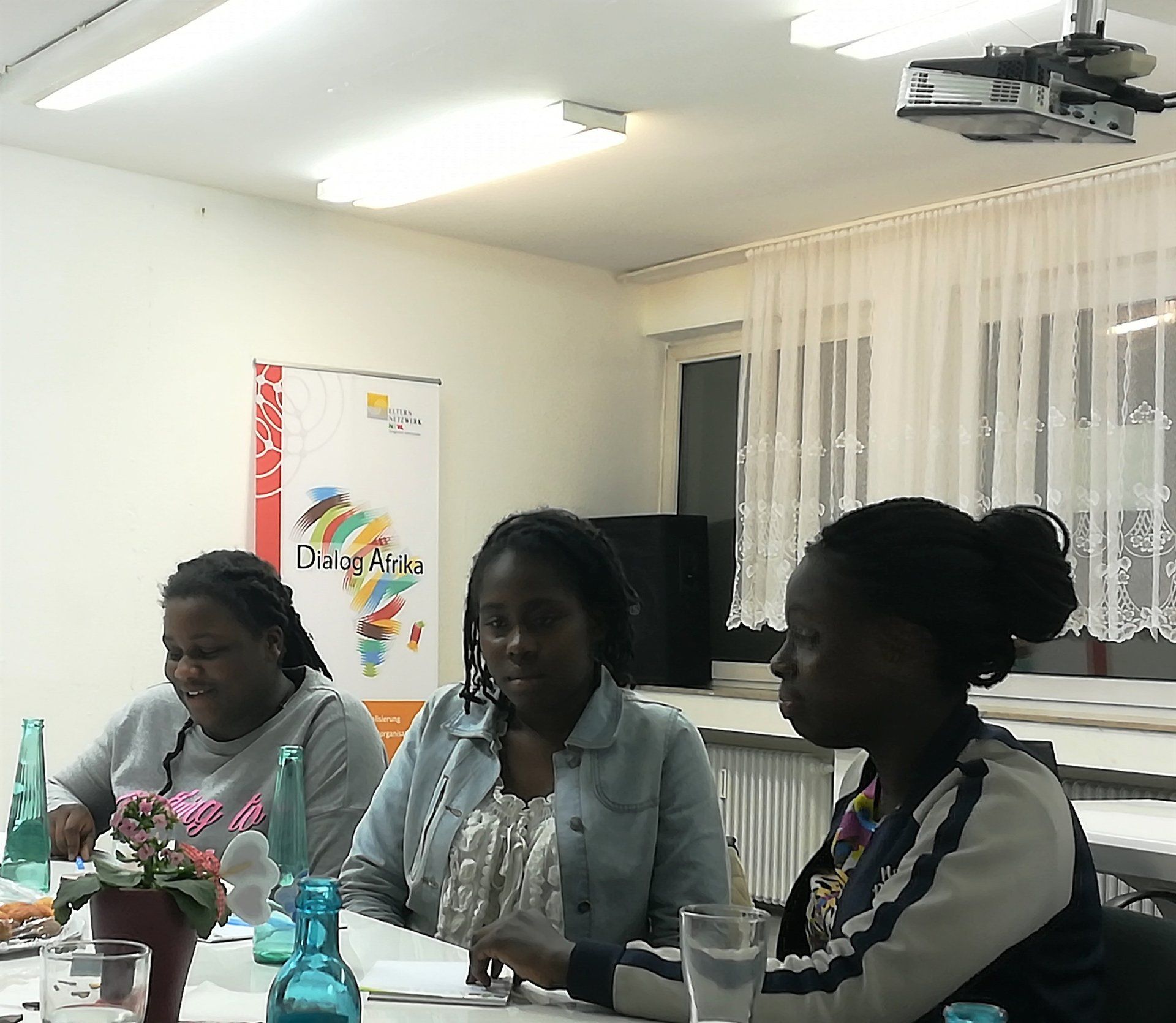 Drei schwarze Frauen an einem Tisch, im Hintergrund des Raumes ein Banner des Projekts Dialog Afrika