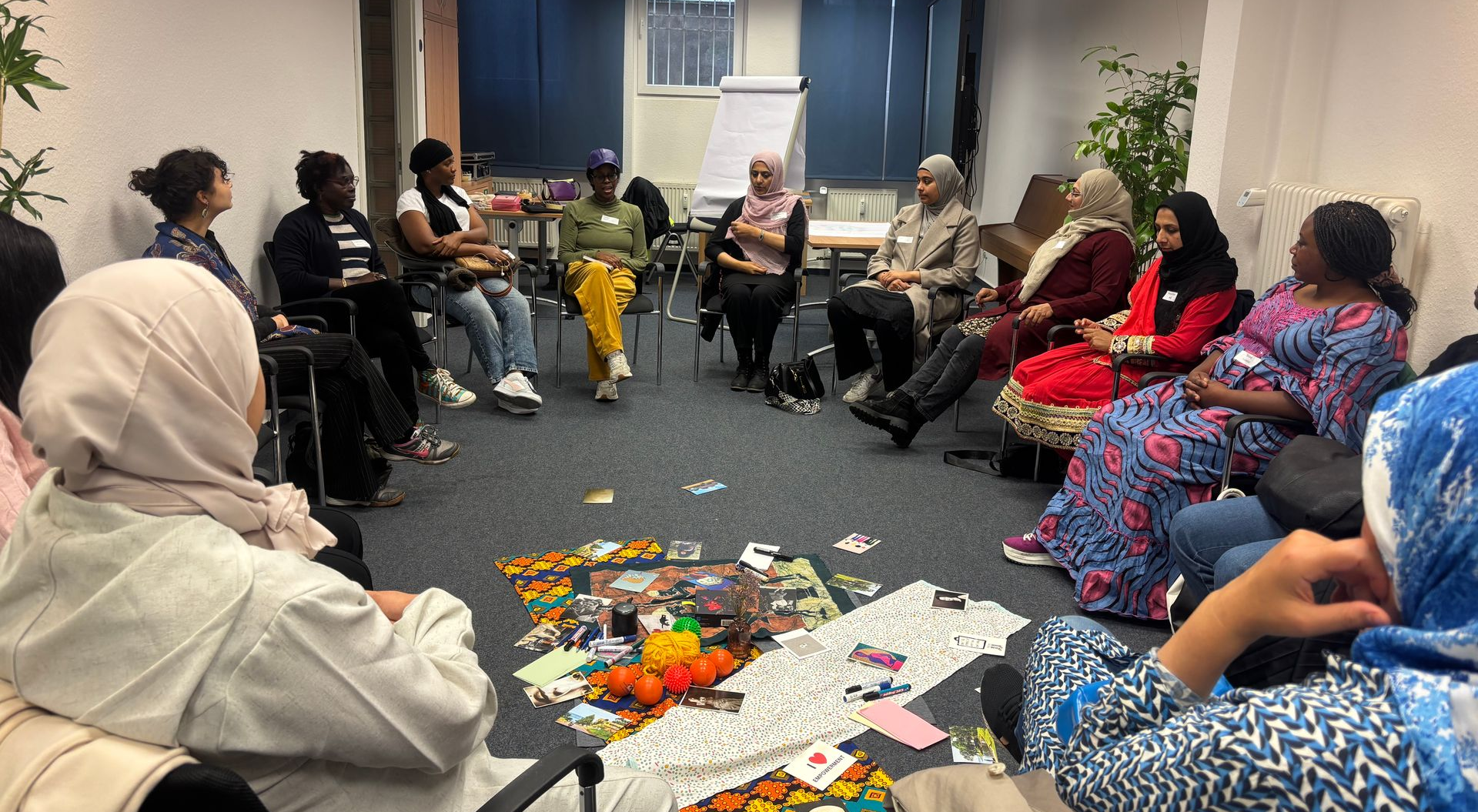 Her-Diversity Frauen in einem Stuhlkreis. In der Mitte liegen viele Karten und kleine Gegenstände.
