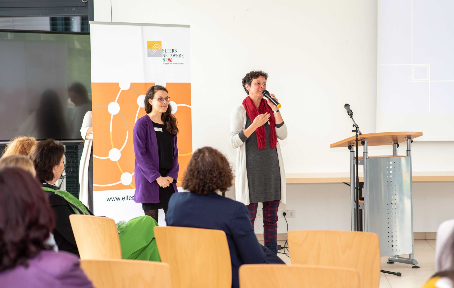 Zwei Sprecherinnen stehen weiter hinten im Raum, eine spricht frei ins Mikrofon. Im Vordergrund das Publikum.