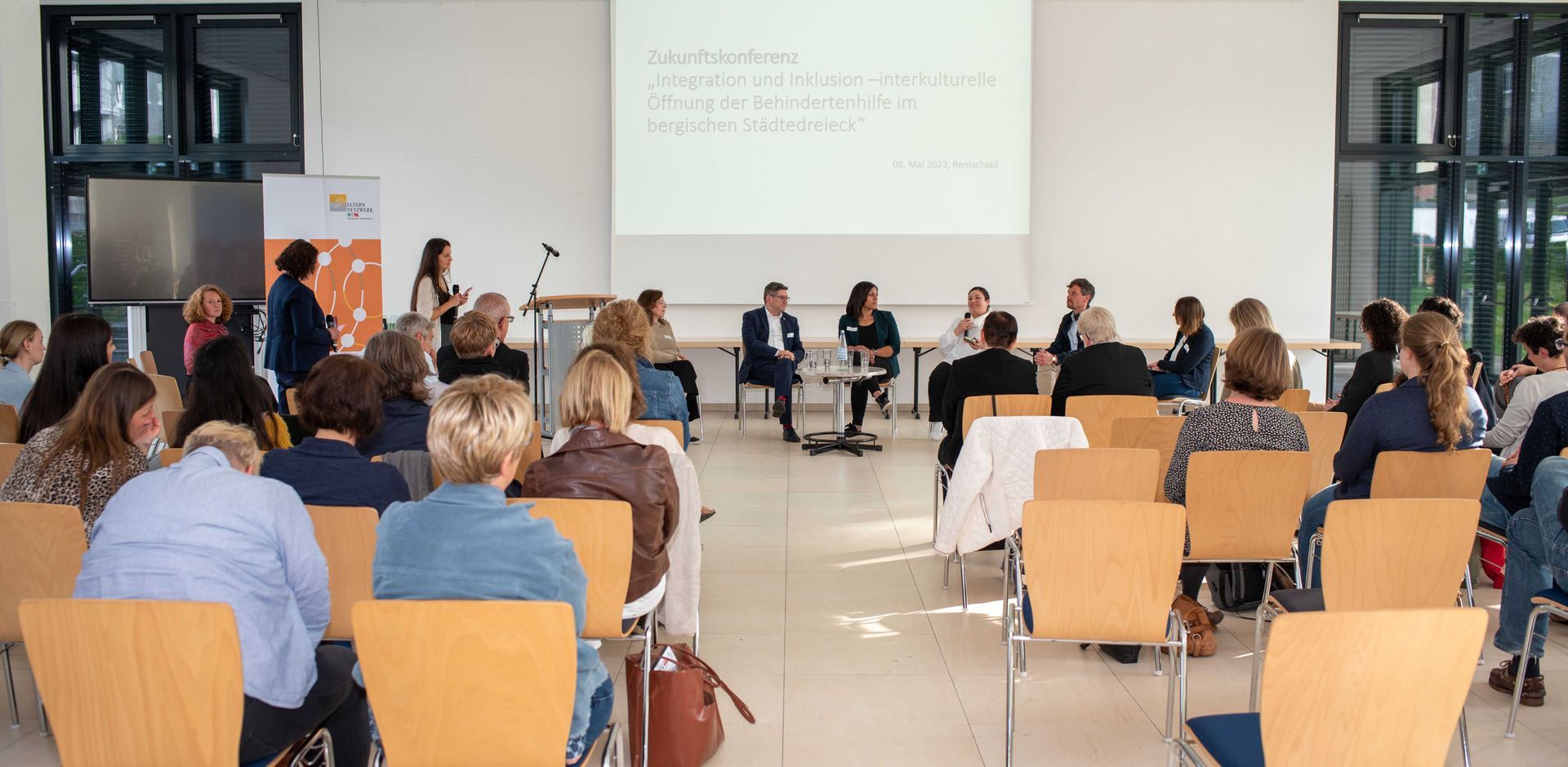 Im Hintergrund, vor der Leinwand, sitzen fünf sich unterhaltende Personen. Im Vordergrund das Publikum der Zukunftskonferenz