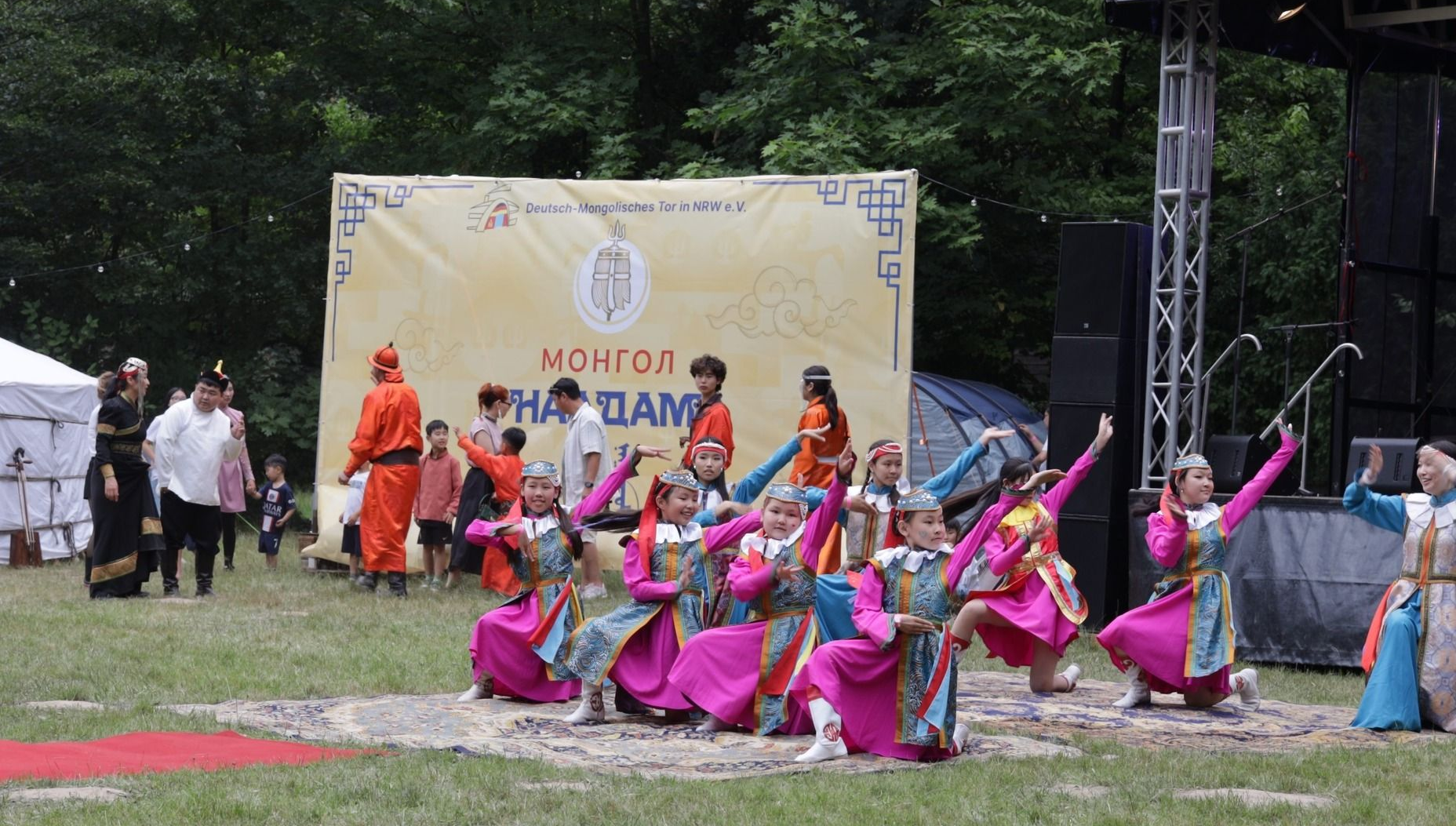 Kinder in traditionellen und farbenfrohen mongolischen Trachten tanzen auf einer Bühne im Park.