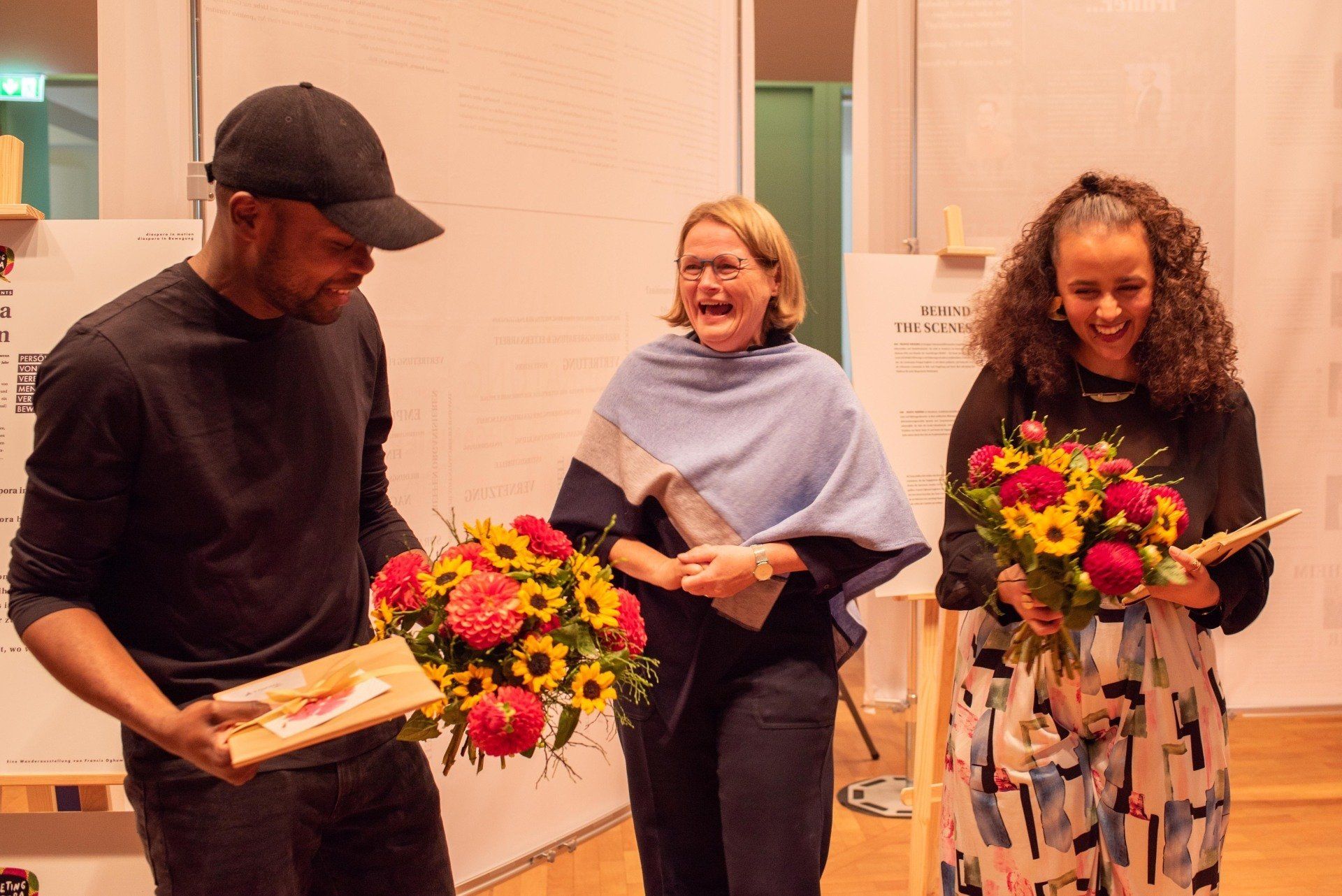 Shavu Nsenga und Francis Oghuma haben gerade u.a. einen großen Blumenstrauß erhalten. Die drei Personen lachen herzlich