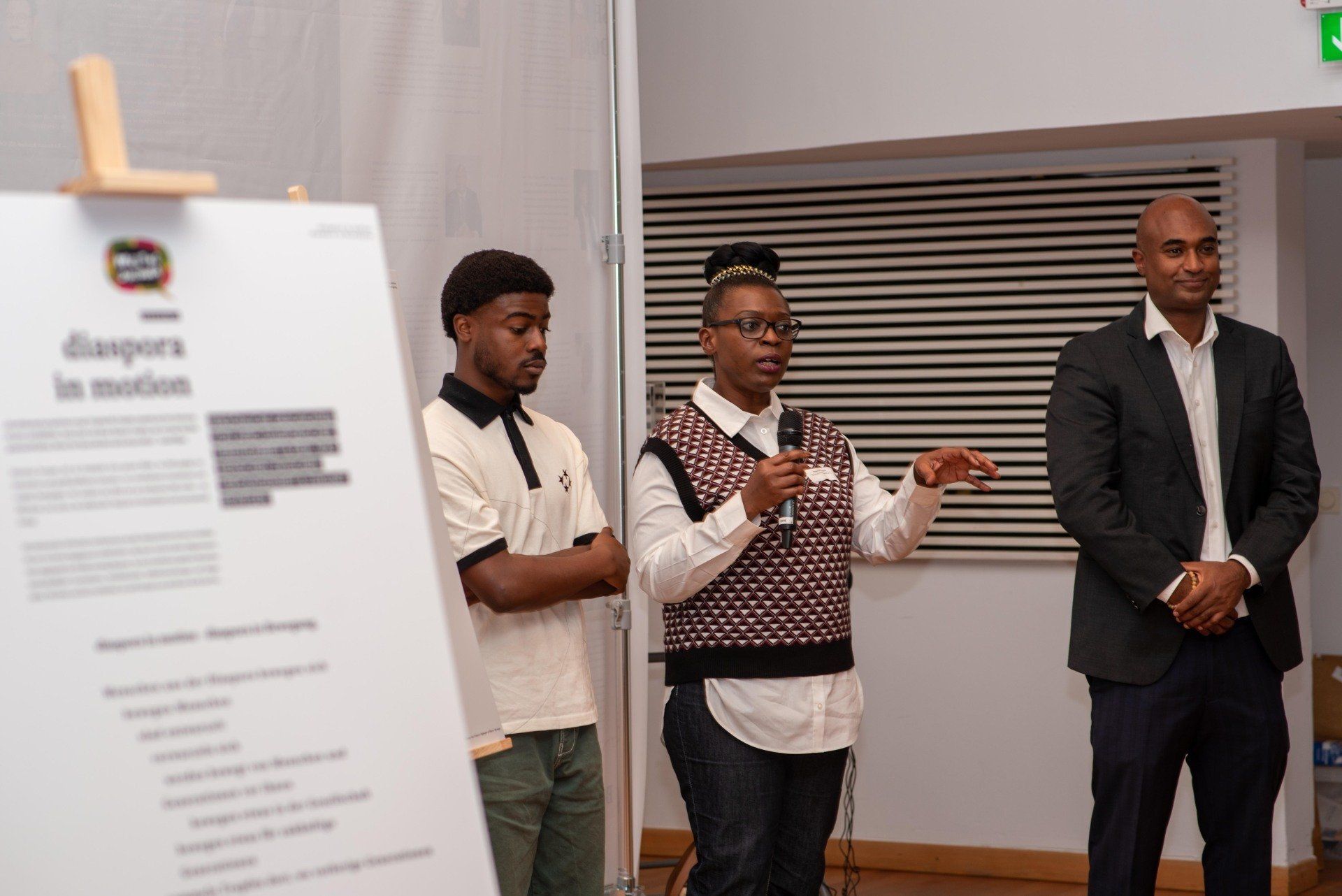 Diaspora in Motion, Ausstellungseröffnung. Im Vordergrund verschwommen eine Informationstafel. Im Hintergrund Besucher*innen