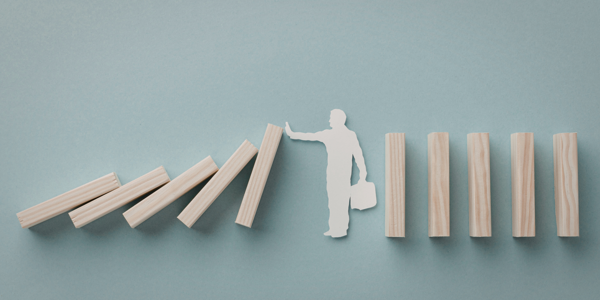 Holz-Dominosteine kippen, eine Person-Silhouette stoppt die Kettenreaktion mit ausgestreckter Hand.