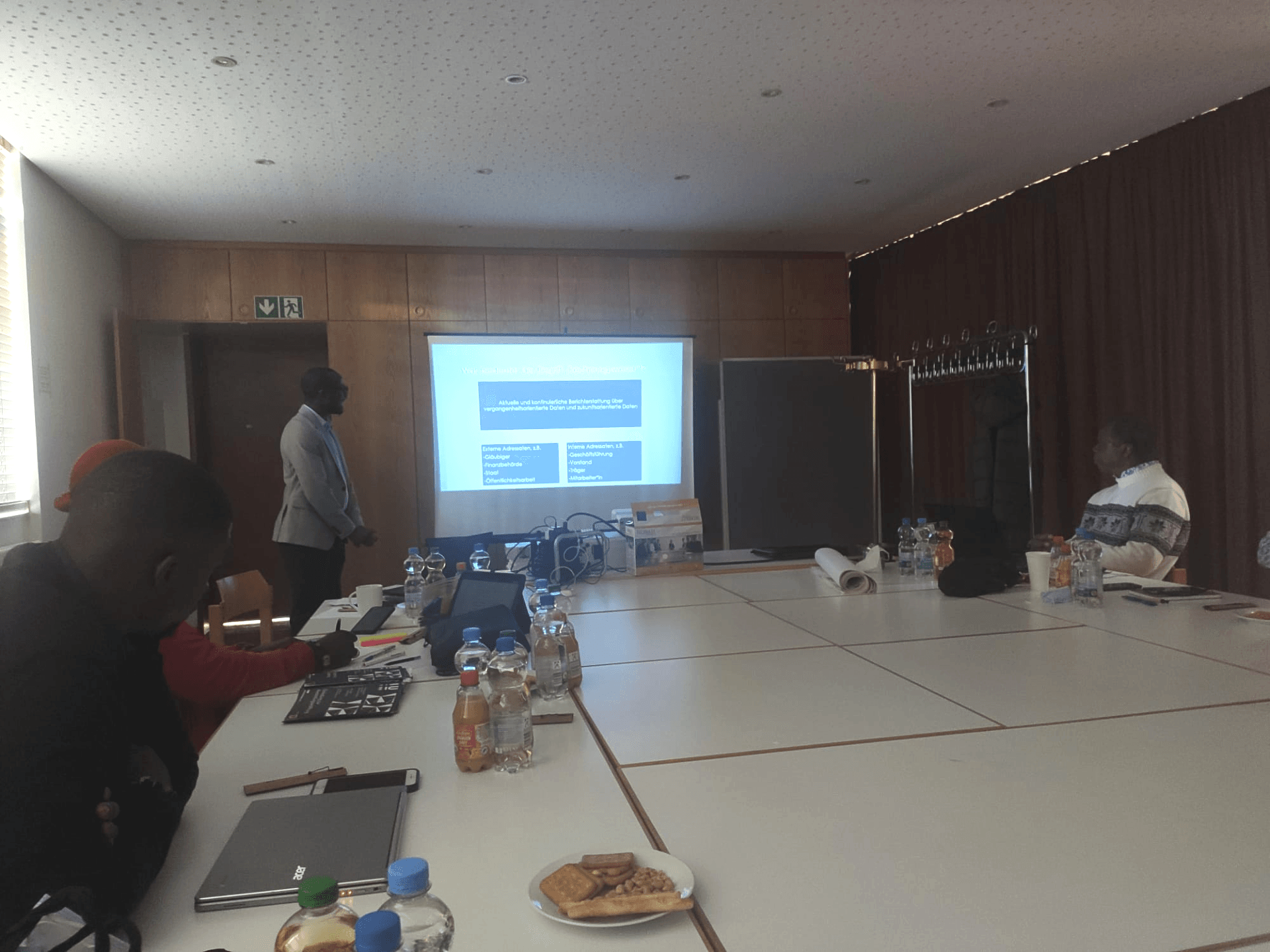 Beim Coaching zum Thema Finanzen sitzen mehre Teilnehmende im Seminarraum. Referent steht vor der Leinwand mit Präsentation