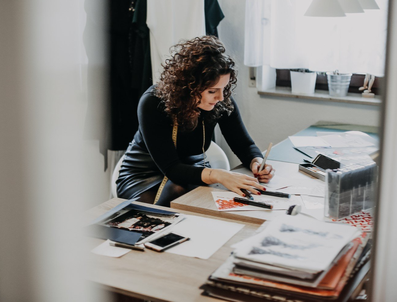 Eine Frau mit langen dunklen, lockigen Haaren sitzt an einem großen Tisch und zeichnet Brautkleider