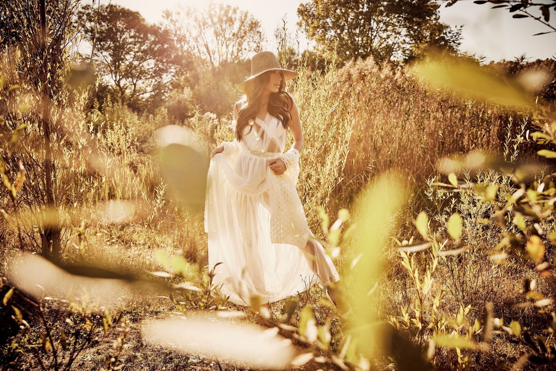 Eine Frau mit langen dunklen Haaren mit einem Hut und einem luftig leichtem Brautkleid steht in einer herbstlich goldenen Natur. Sie blickt vertraeumt zur Seite.