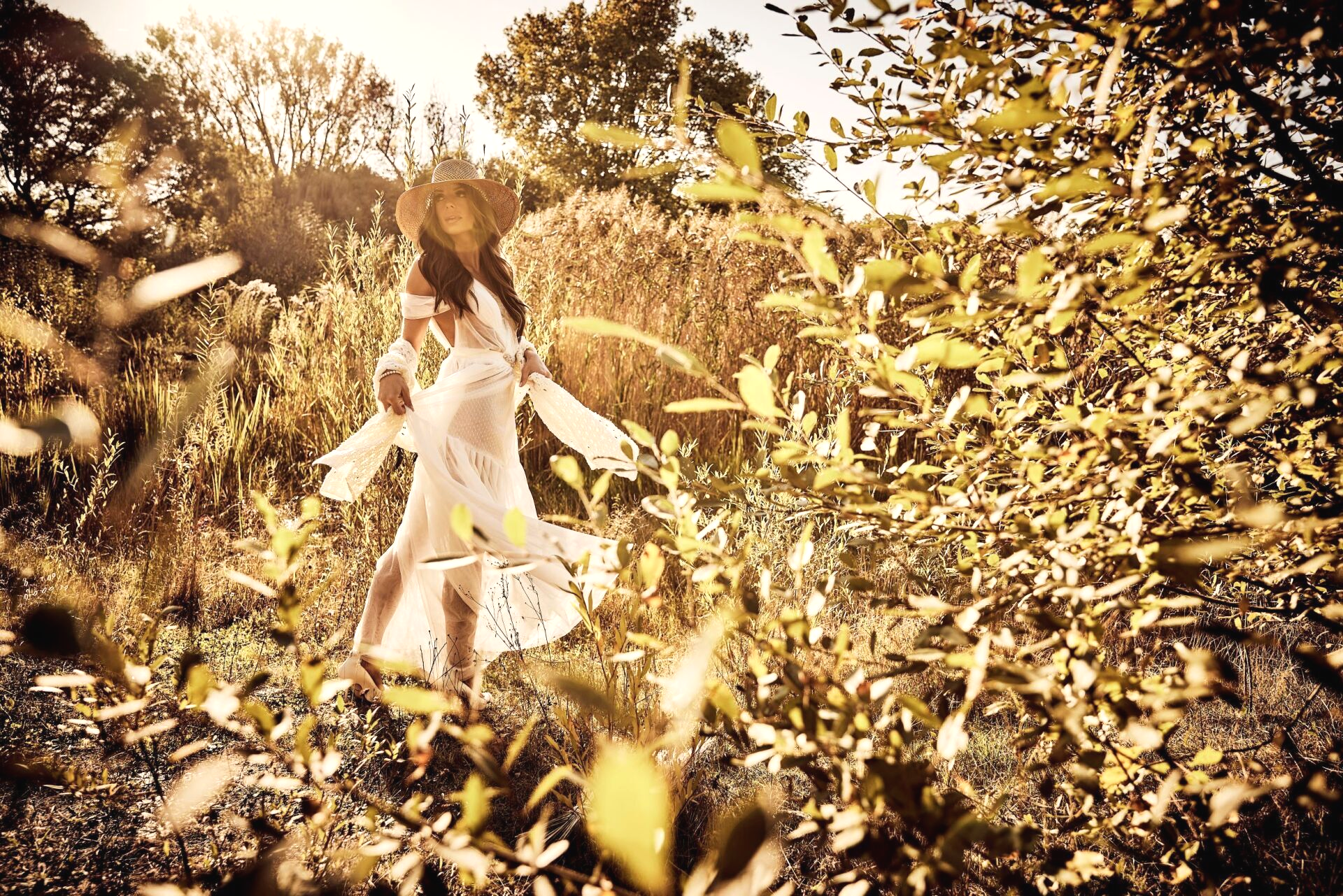 Eine Frau in einem luftig leichtem Brautkleid steht mit dem Rücken zum Betrachter in einer herbstlich, goldenen Natur. Sie trägt einen Hut und dreht sich in einer schwungvollen Drehung im Oberkörper zum Betrachter.