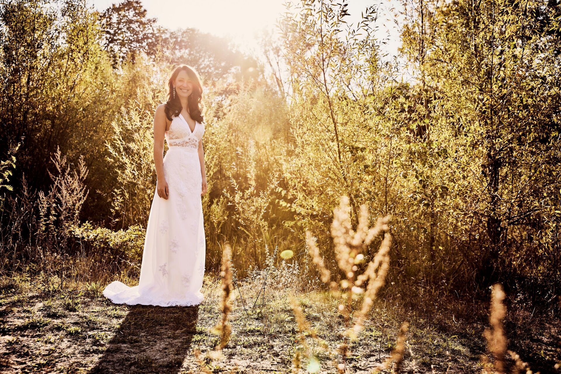 Eine Frau mit langen dunklen Haaren in einem besticktem Brautkleid steht am linken Bildrand in einer herbstlich goldenen Natur. Sie laechelt den Betrachter an.