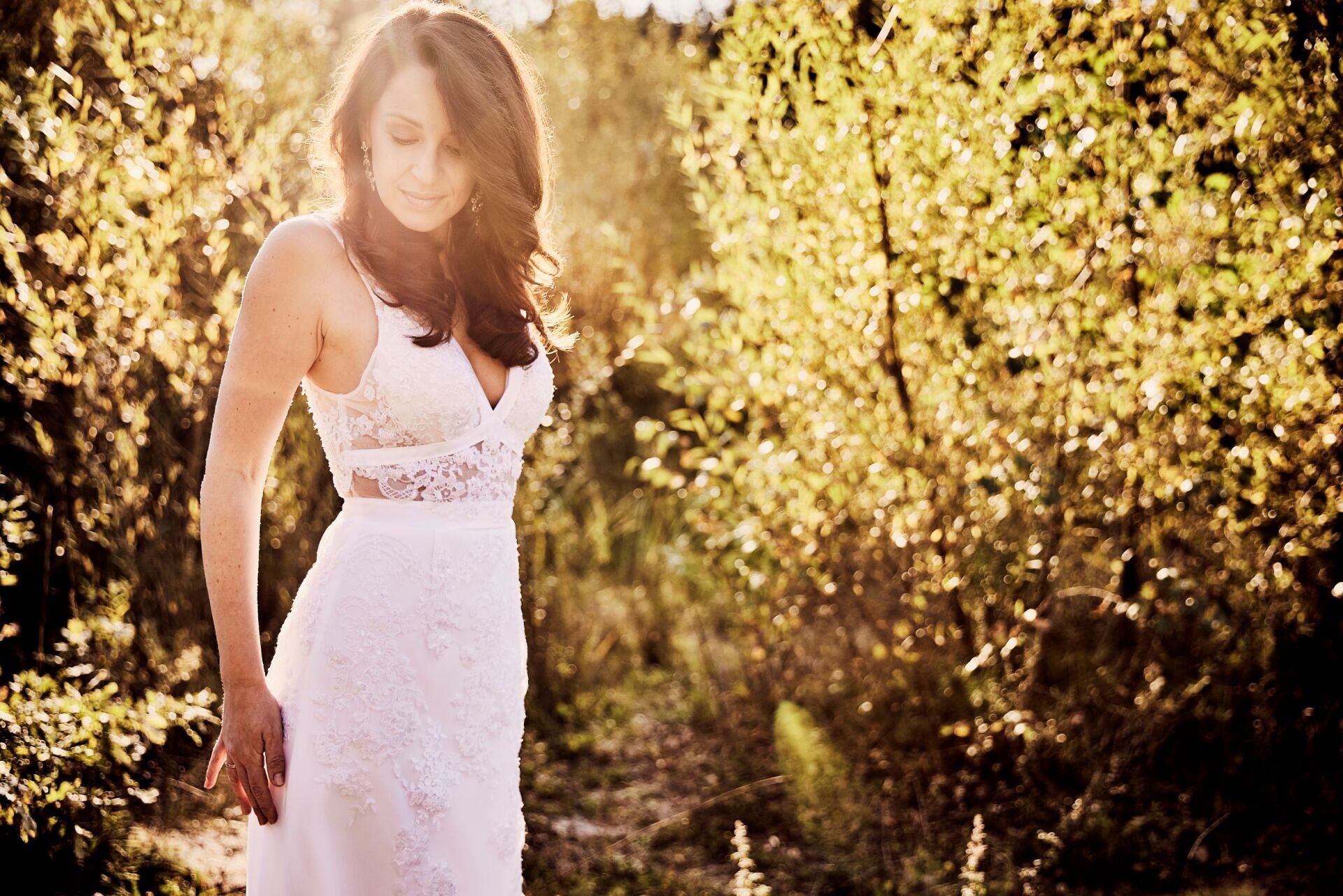 Eine Frau mit langen dunklen Haaren in einem besticktem Brautkleid steht am linken Bildrand in einer herbstlich goldenen Natur. Das Bild ist angeschnitten und zeigt nur den Oberkoerper. Sie laechelt und sieht zufrieden an sich herab.