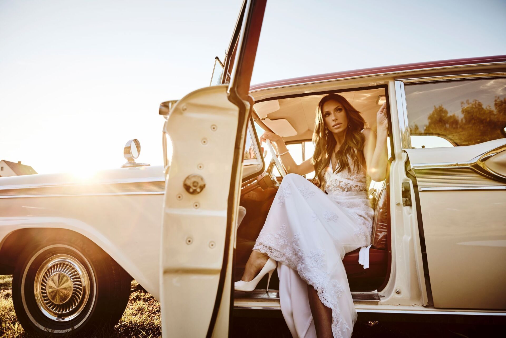 Eine Frau mit langen dunklen Haaren in einem Spitzenbestickten Brautkleid steigt aus einem cremefarbenen Oldtimers aus.