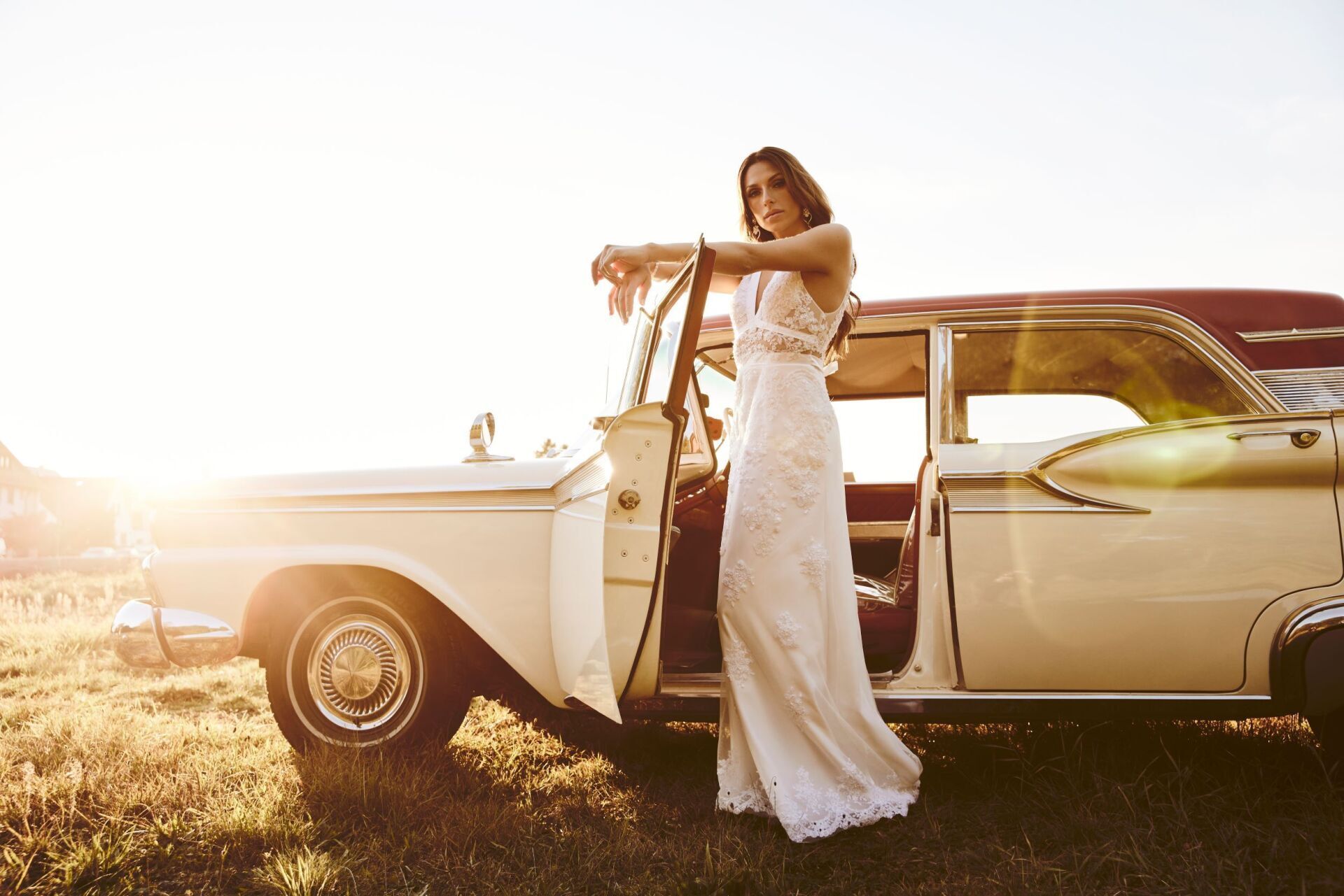 Eine Frau mit langen dunklen Haaren steht mit einem Spitzenbestickten Brautkleid an der offenen Tuer eines cremefarbenen Oldtimers.