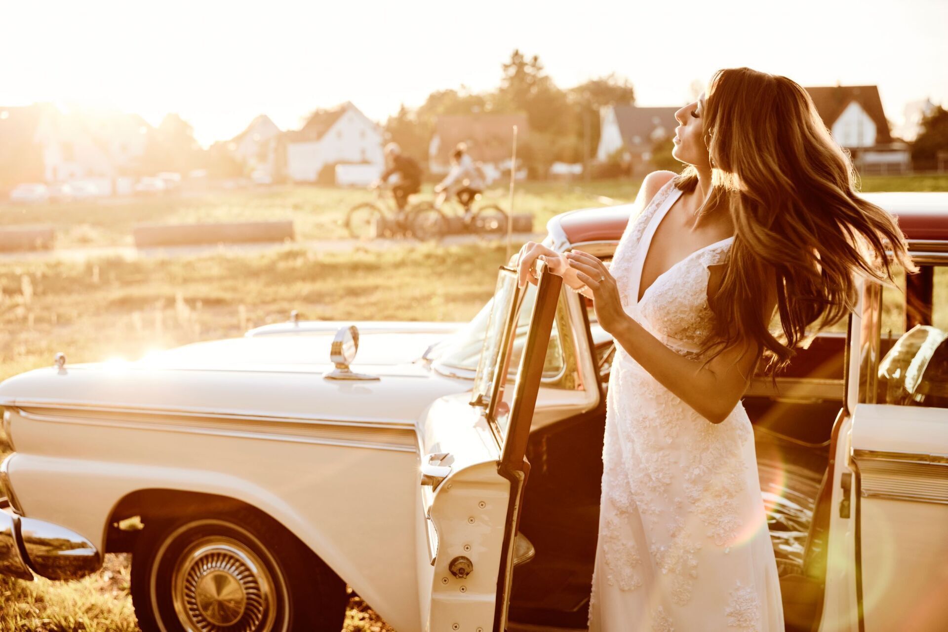 Eine Frau mit langen dunklen Haaren mit einem Spitzenbestickten Brautkleid steht an der offenen Tür eines cremefarbenen Oldtimers und schwingt ihre Haare.