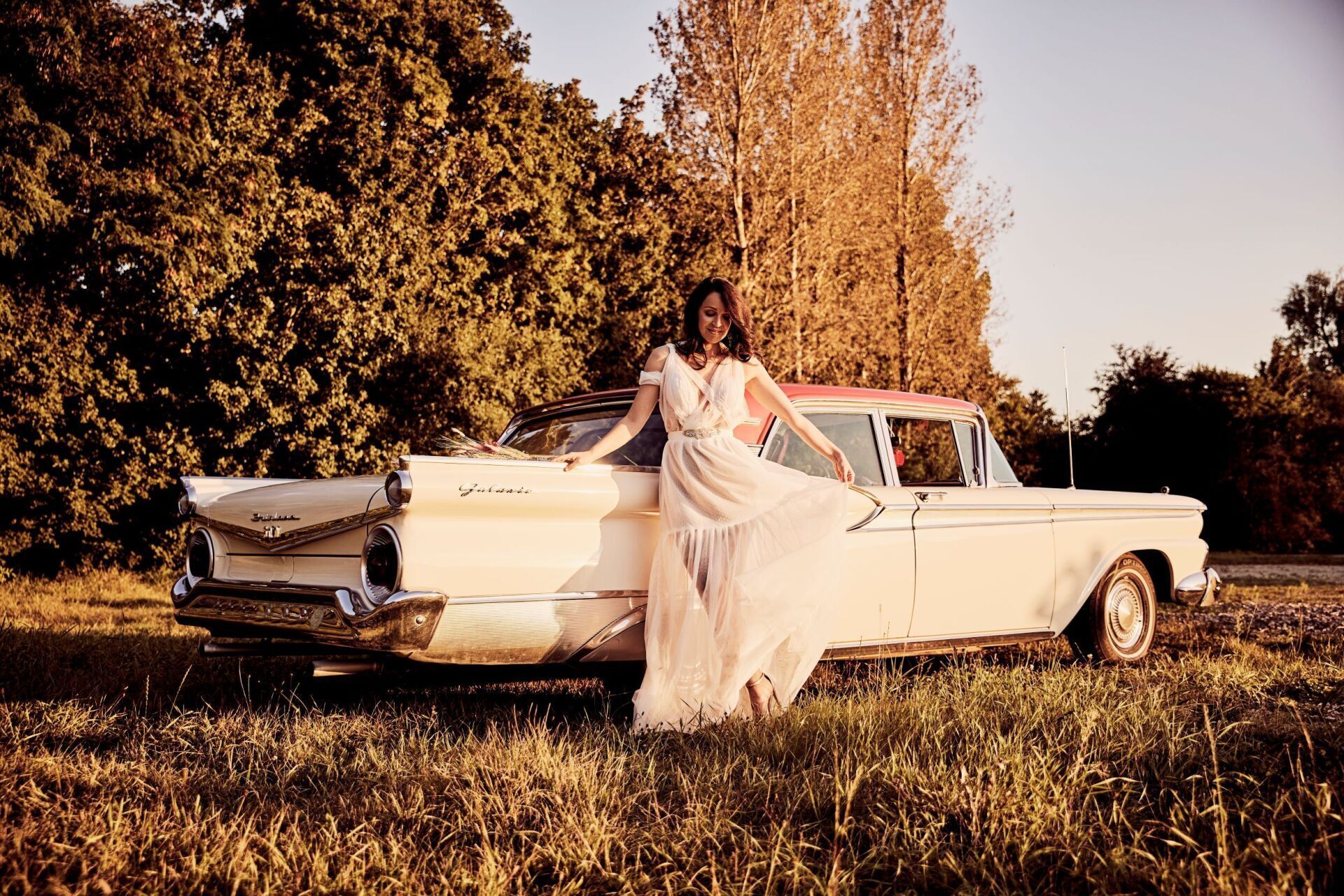 Eine Frau mit langen dunklen Haaren und einem luftig leichtem Brautkleid steht vor einem cremefarbenem Oldtimer und schwingt mit dem Kleid.