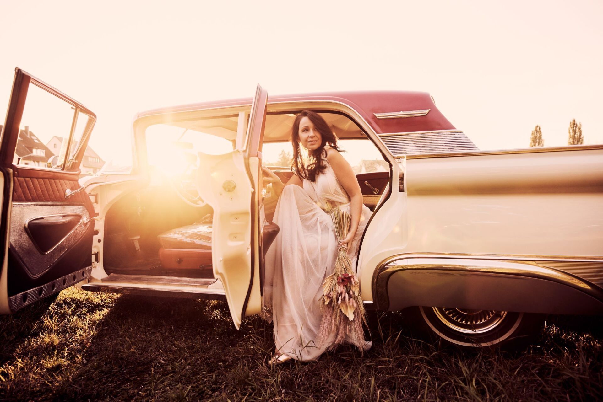Eine Frau mit langen dunklen Haaren und einem luftig leichtem Brautkleid steigt gerade aus einem cremefarbenem Oldtimer aus.