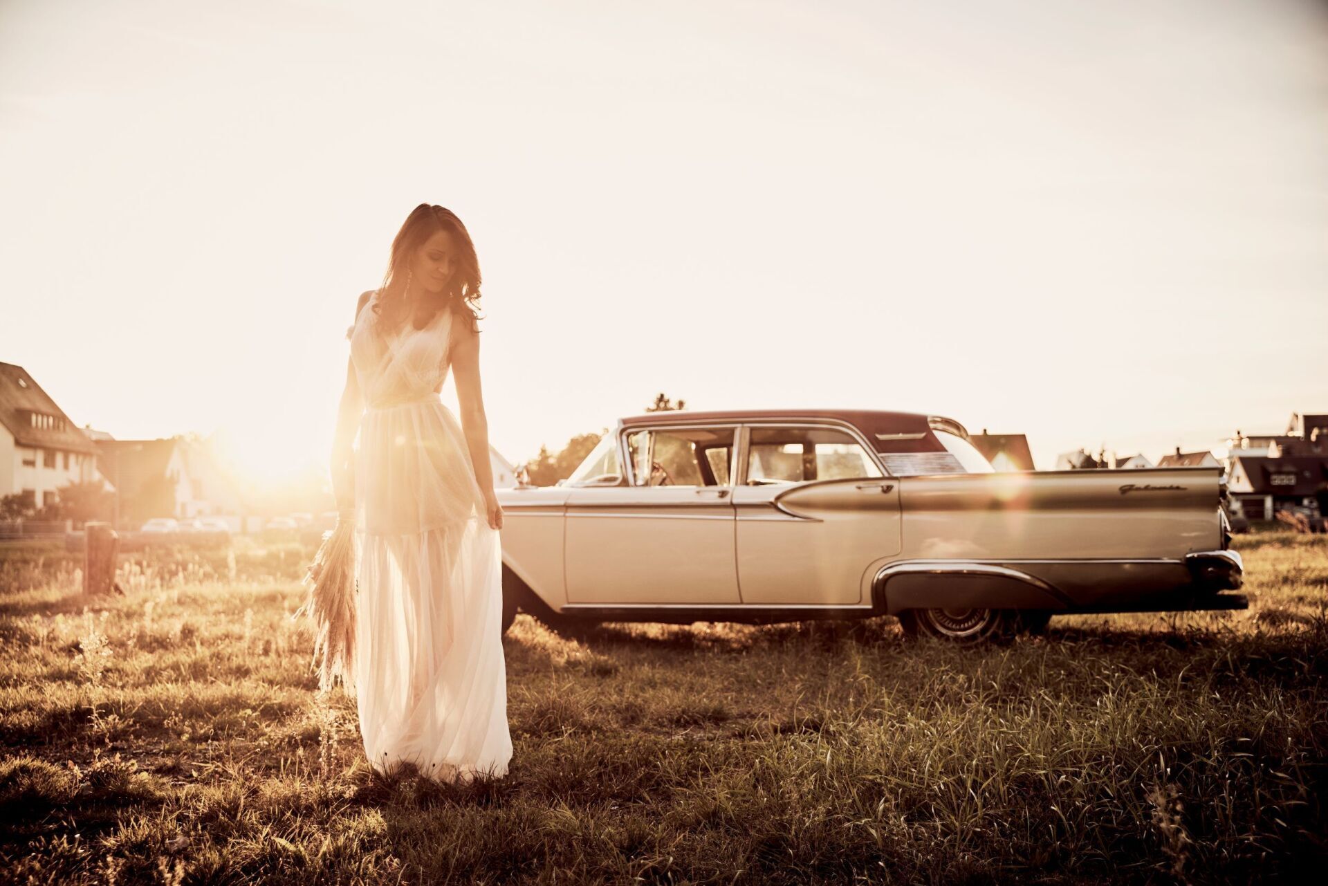 Eine Frau mit langen dunklen Haaren und einem luftig leichtem Brautkleid steht in der linken Bildhaelfte vor einem cremefarbenem Oldtimer. Sie blickt vertraeumt zur Seite.