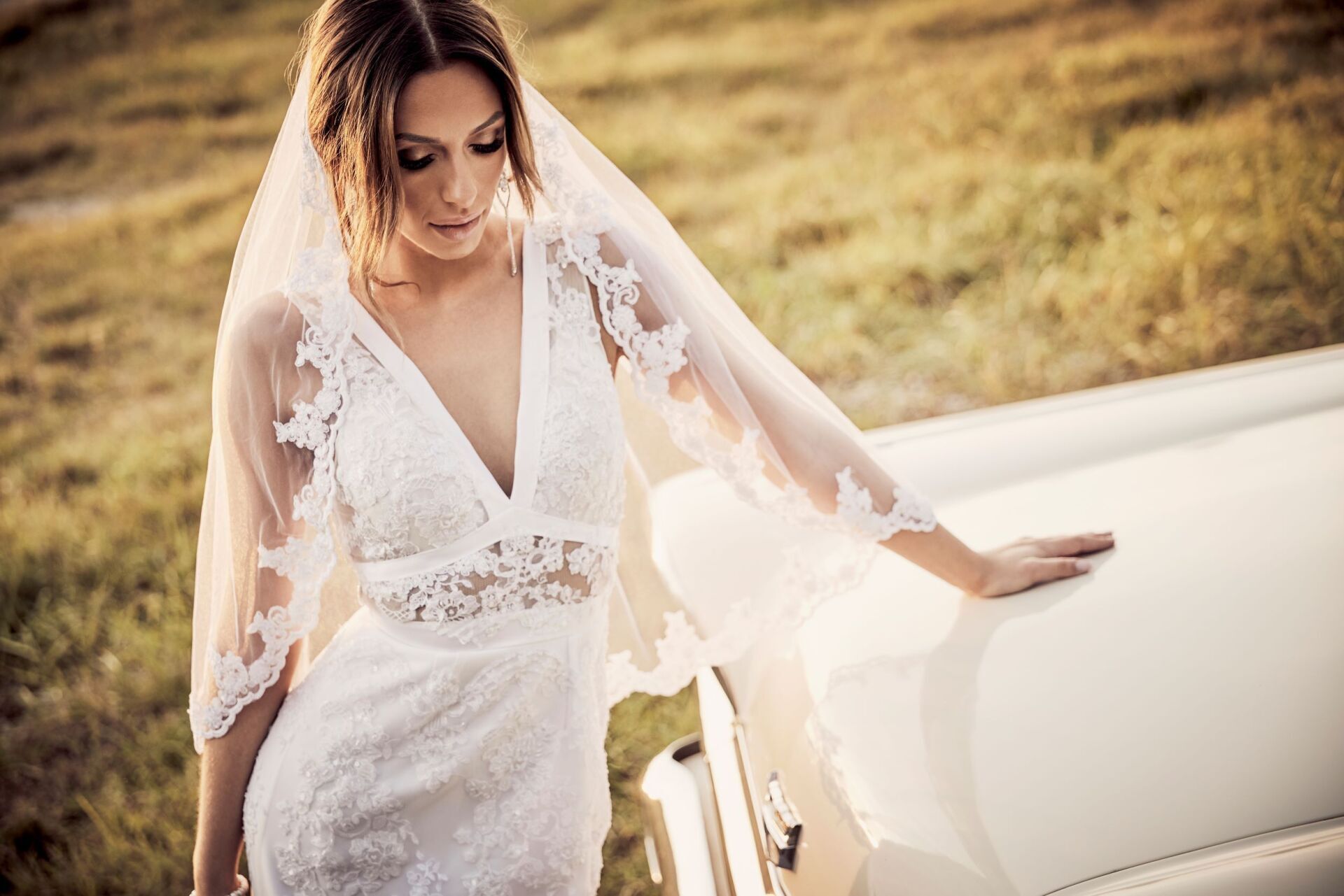 Eine Frau mit langen dunklen Haaren und einem Schleier steht mit einem Spitzenbestickten Brautkleid vor der Motorhaube eines cremefarbenen Oldtimers. Sie blickt zu Boden.