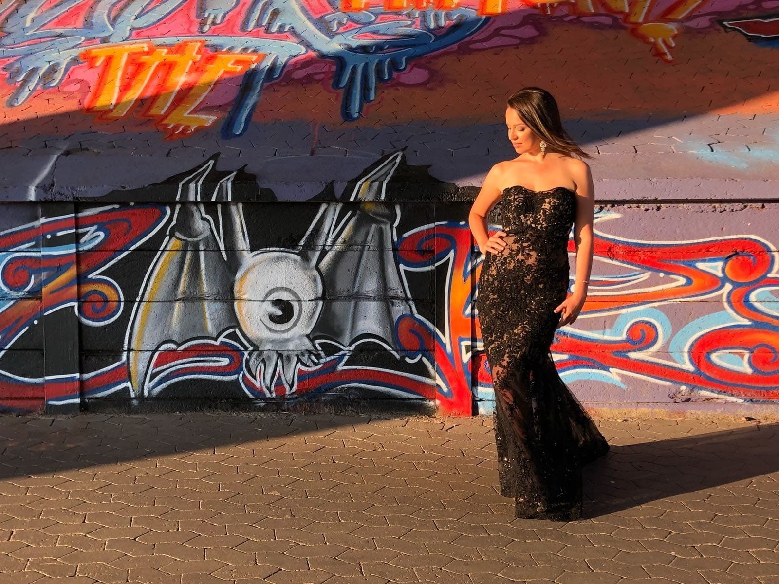 Eine Frau mit langen dunklen Haaren, steht vor einem großen bunten Graffiti. Sie traegt ein langes, teilweise durchsichtiges Spitzenkleid in schwarz. Sie blickt seitlich aus dem Bild heraus.