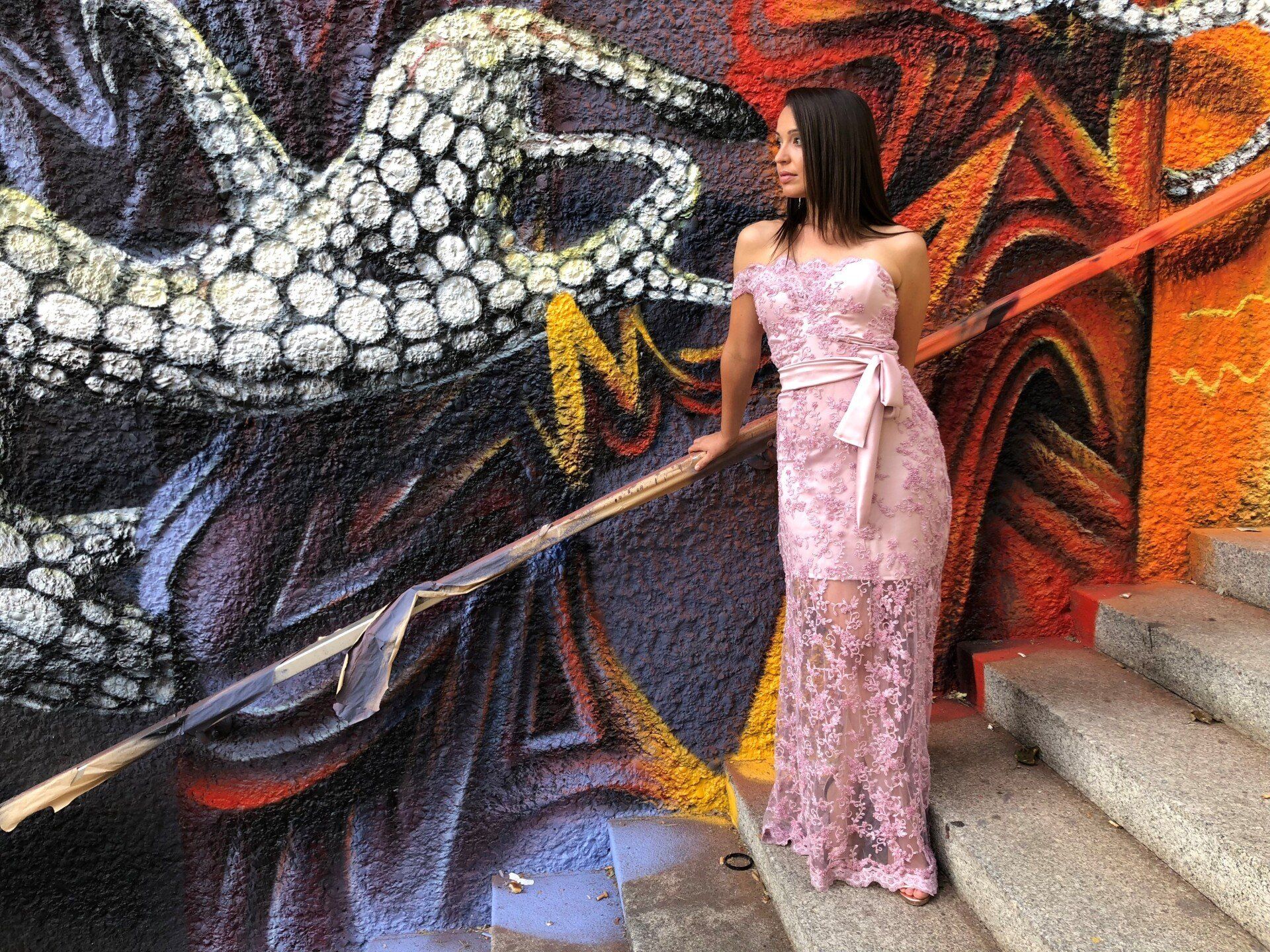 Eine Frau mit langen dunklen Haaren, steht auf einer Treppe vor einem großen bunten Graffiti. Sie traegt ein langes, teilweise durchsichtiges Spitzenkleid in rosa. Sie blickt seitlich aus dem Bild heraus.