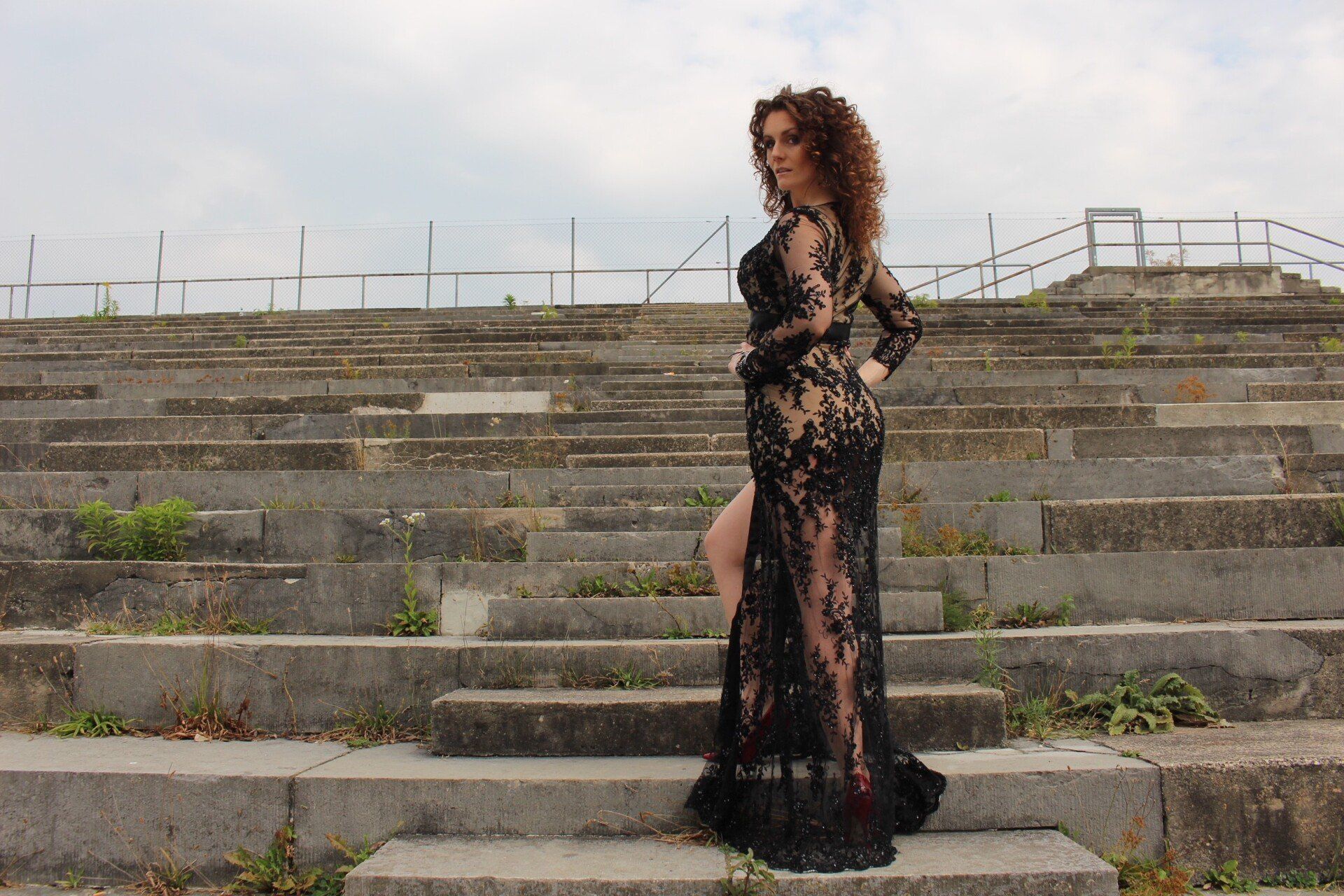 Eine Frau mit dunkelblonden Locken traegt ein langaermeliges, durchsichtiges Spitzenkleid auf Steinstufen. Sie steht mit dem Ruecken zum Betrachter dreht aber den Torso zum Betrachter.