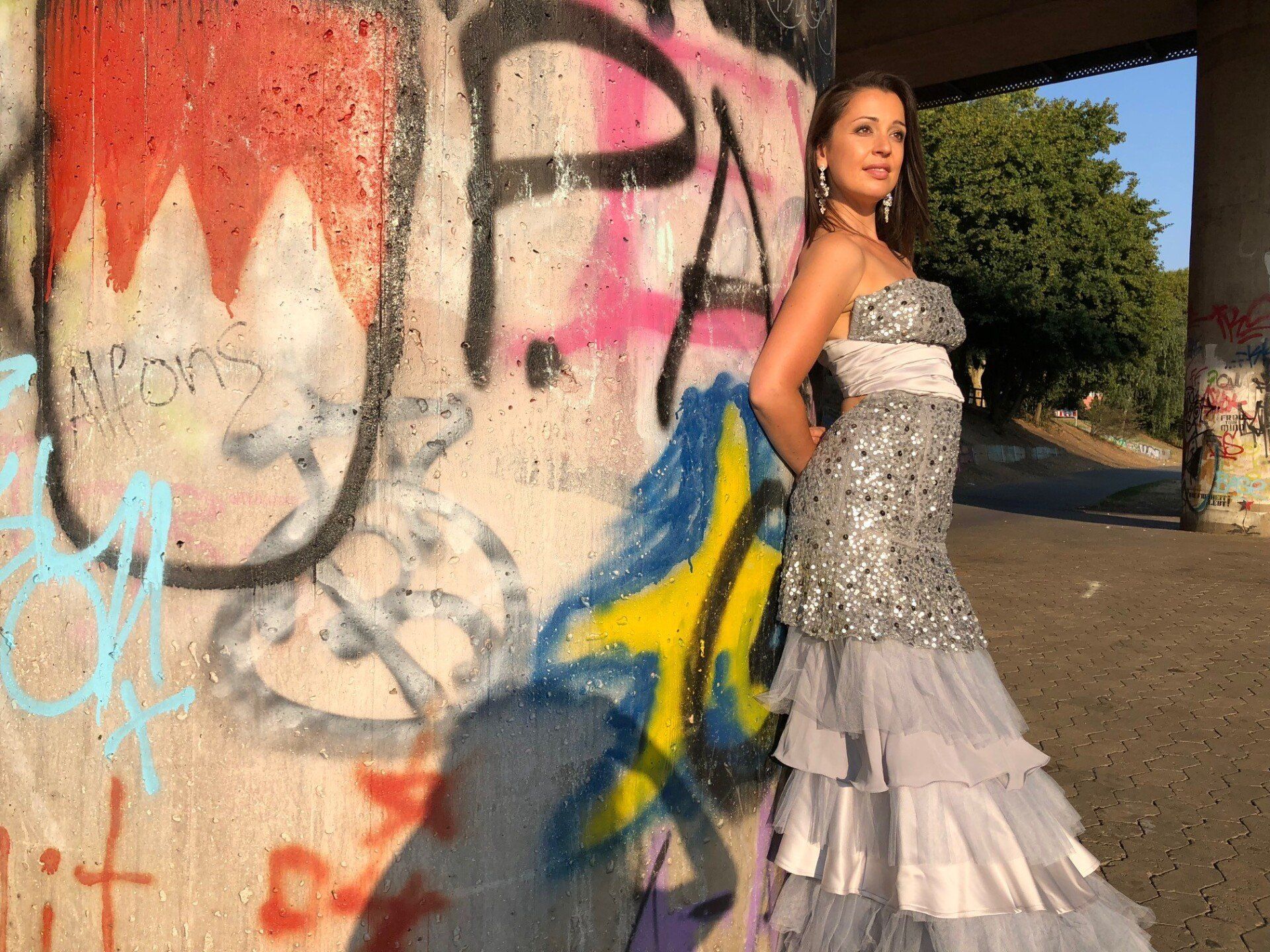 Eine Frau mit langen dunklen Haaren, steht vor einer großen Betonsaeule mit bunten Graffiti. Sie traegt ein langes Paillettenbesetzes Kleid in silber. Sie lehnt seitlich an der Saeule und blickt aus dem Bild heraus.