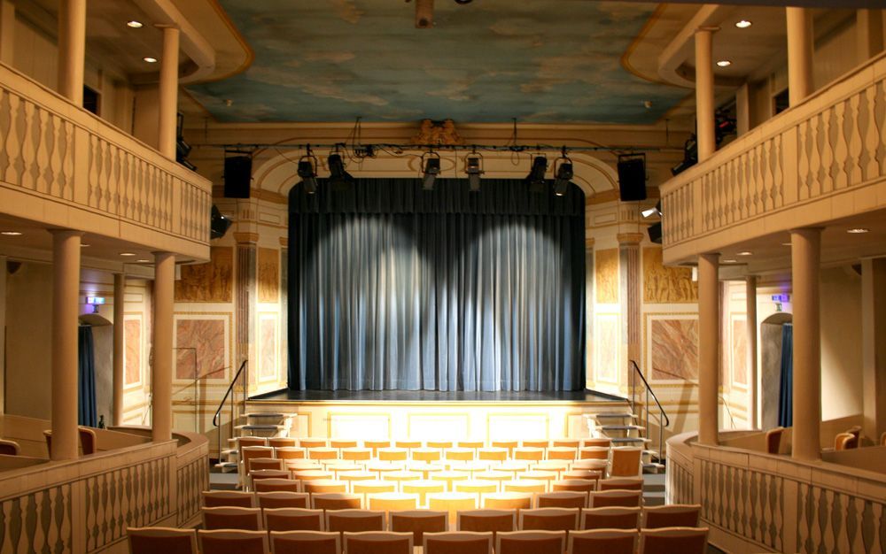 Theatersaal im Comoedienhaus Wilhelmsbad mit Reihenbestuhlung und seitlich die Logen, Blick auf die Bühne mit schwarzem Vorhang
