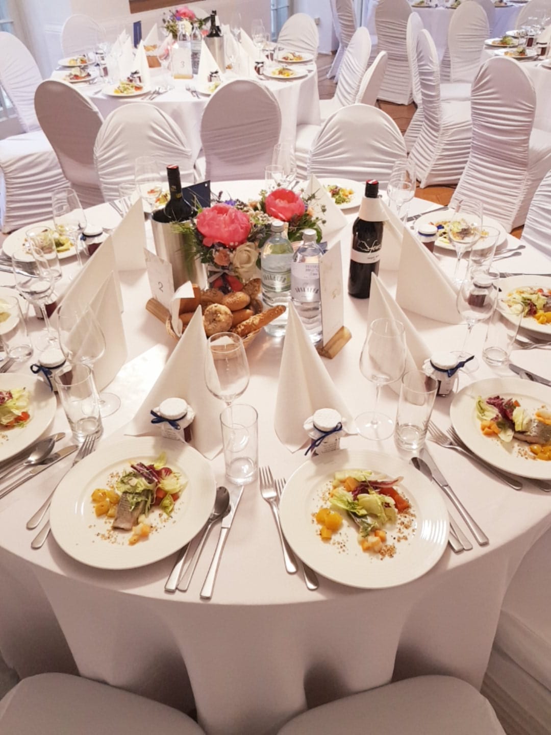 Festlich gedeckter runder Tisch mit weißen Hussen und Blumenschmuck.