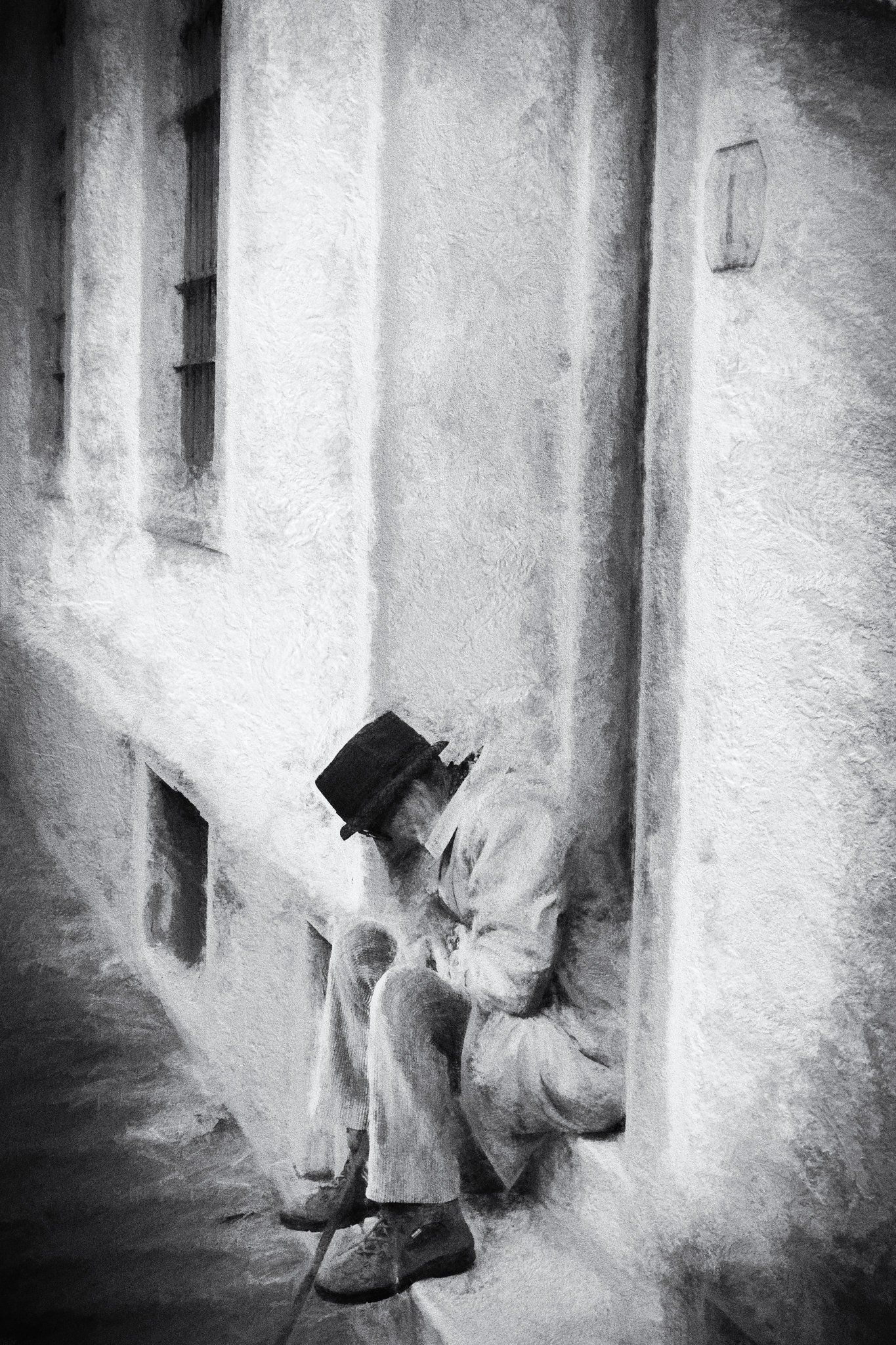 Ein älterer Mann mit Mantel und Hut sitzt zusammengekauert in einem Hauseingang in Florenz. Ein Moment der Stille. Impressionistische Straßenfotografie aus Italien in schwarz weiß.