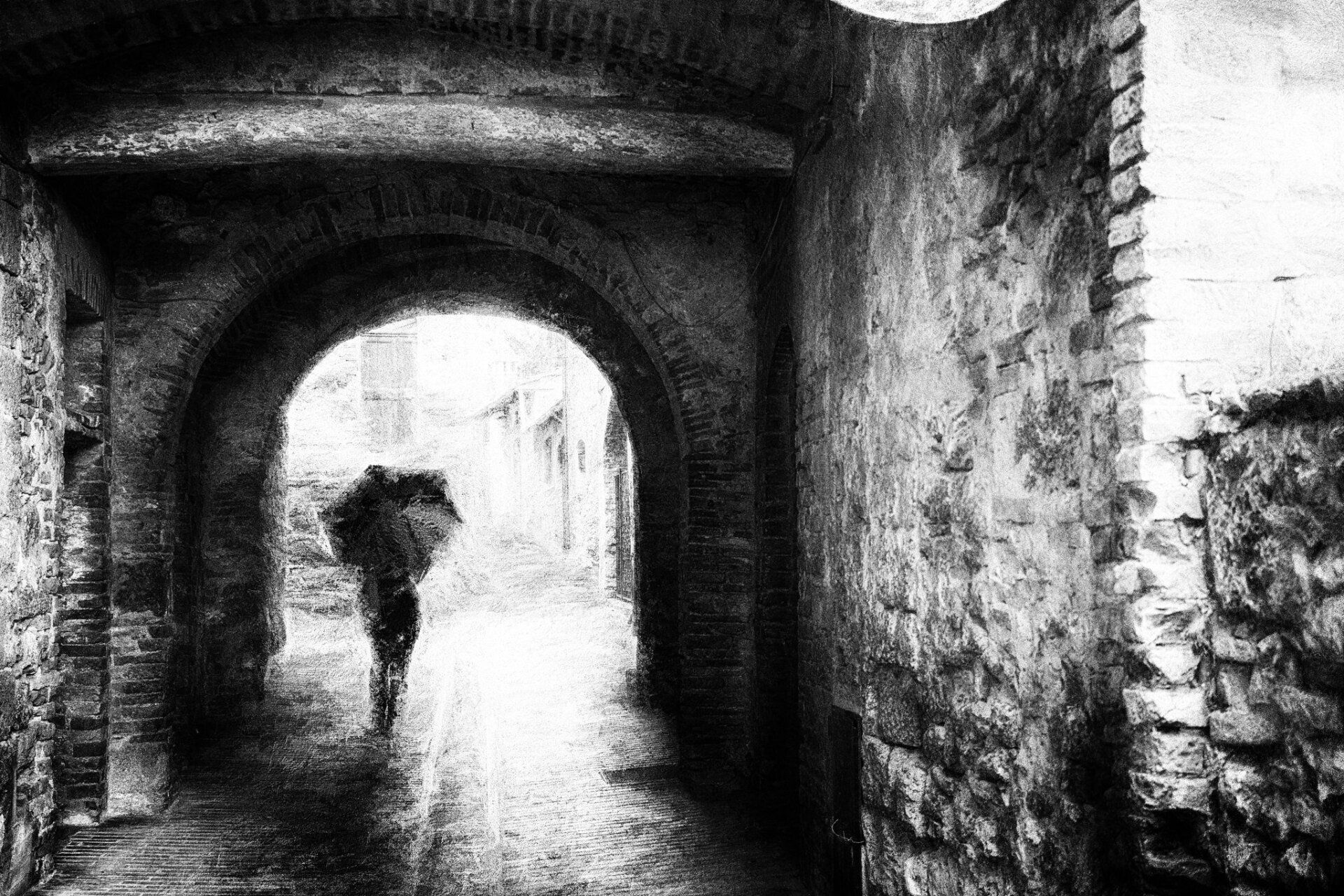 Eine Frau mit Regenschirm steht geht durch einen Hausdurchgang in der Altstadt von San Gimignano Toskana. Impressionistische Straßenfotografie aus Italien in schwarz weiß