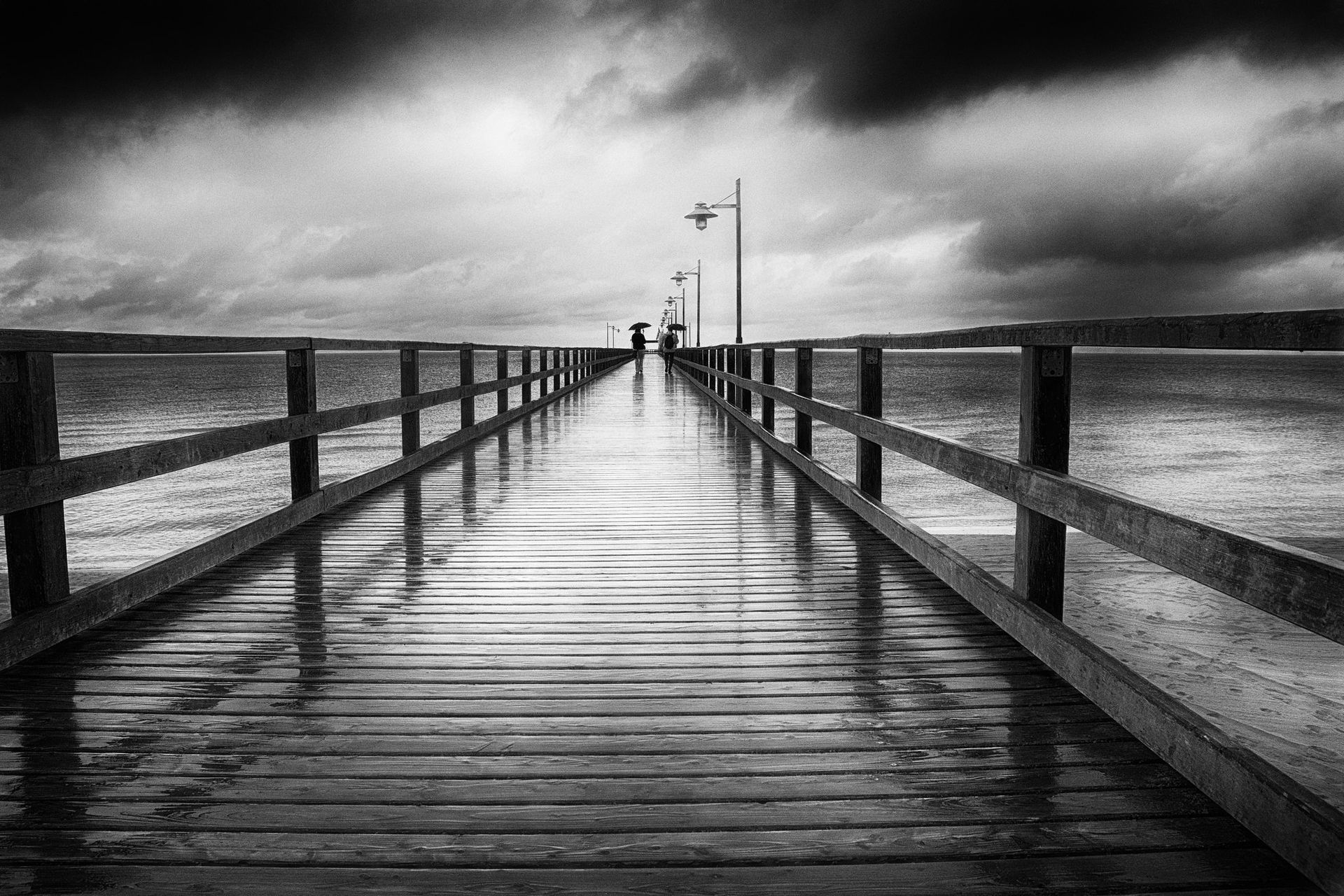 Zwei Menschen mit Regenschirmen stehen auf der Regennassen Seebrücke von Bansin am Strand der Ostsee. Straßenfotografie aus Deutschland in schwarz weiss