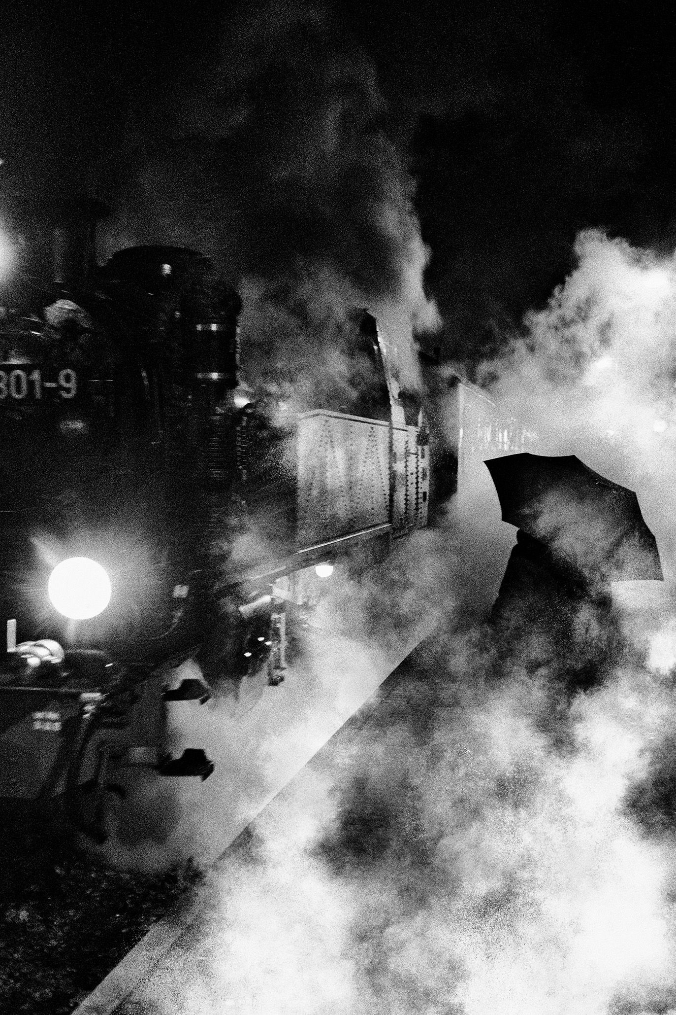 Schwarz weiß Fotografie einer Frau mit Regenschirm neben einer qualmenden Dampflokomotive