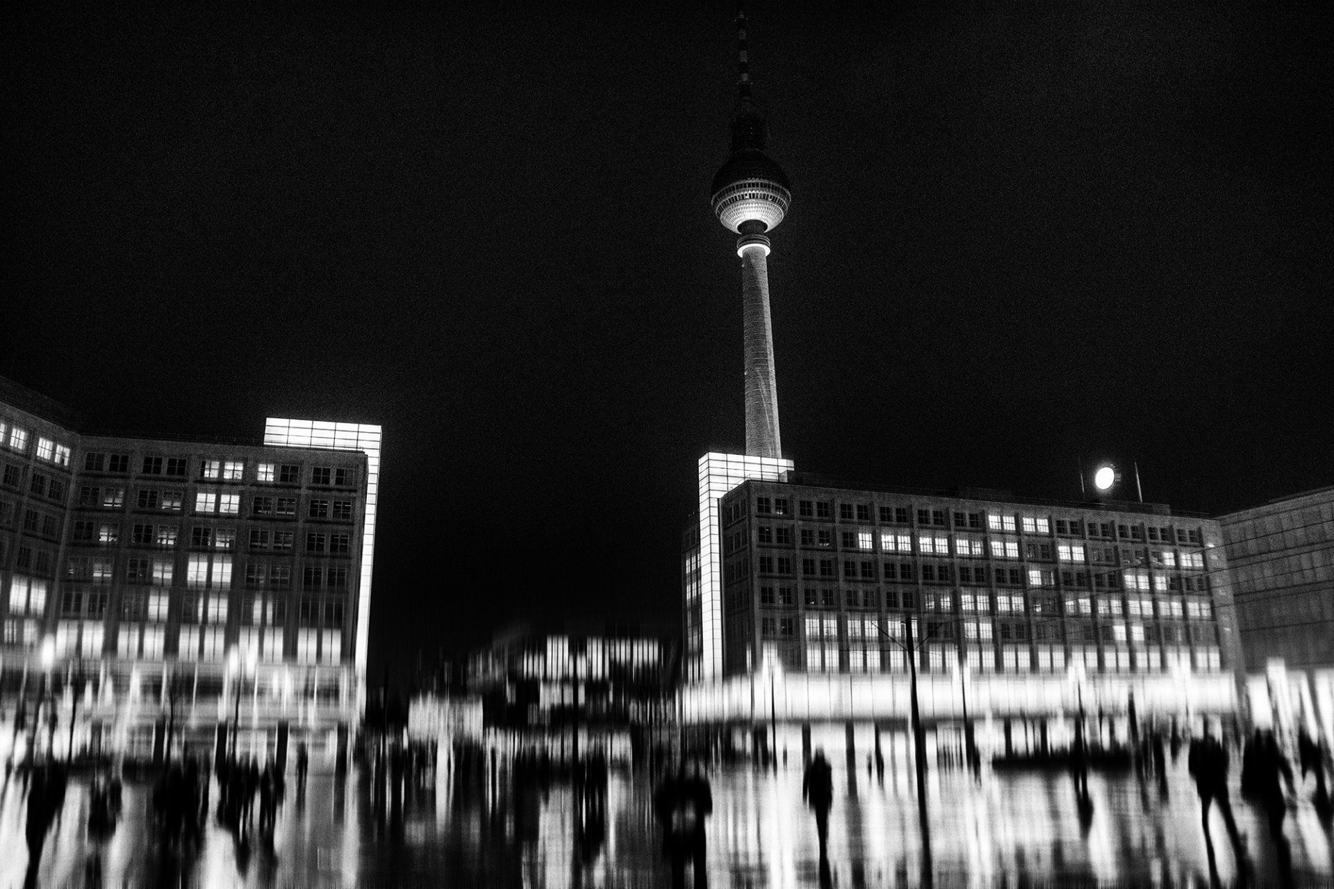 Eine regnerische Nacht in Berlin. Menschen gehen über den nassen beleuchteten Alexanderplatz in der Mitte Berlins. Nachtfotografie aus Berlin in schwarz und weiß.