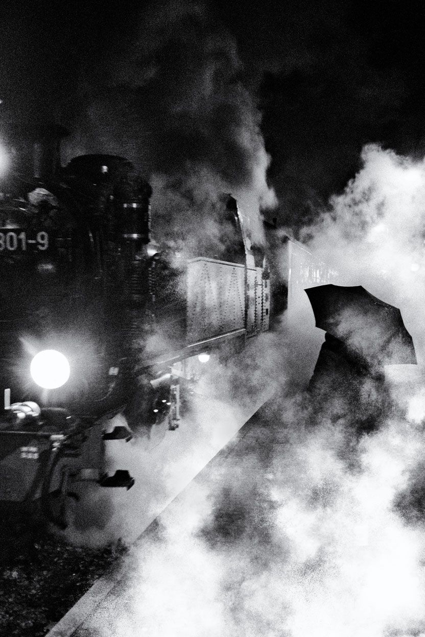 Eine Frau mit Regenschirm steht neben einer Dampflokomotive auf dem Bahnsteig. Straßenfotografie aus Deutschland in schwarz weiss