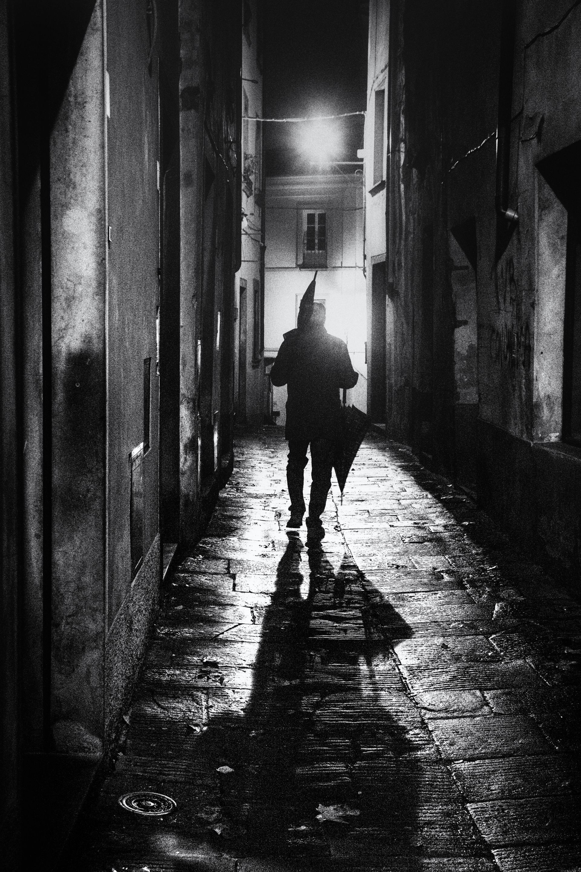 Ein Mann mit zwei Regenschirmen geht durch eine enge Gasse in der Altstadt von Barga. Die Straßenbeleuchtung wirft einen langen Schatten auf dem regennassen Straßenpflaster. Straßenfotografie aus Italien in schwarz weiss.