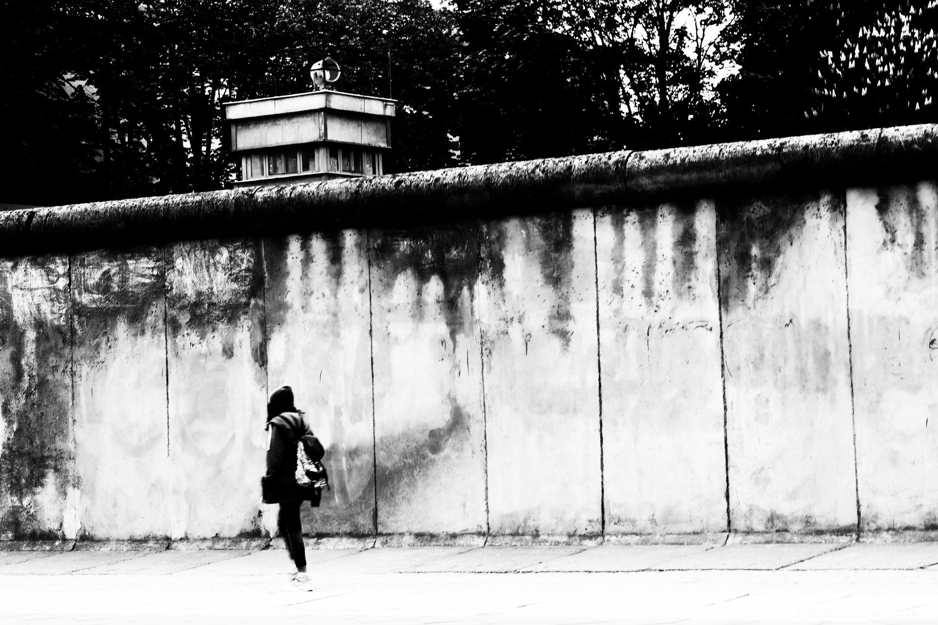Schwarz weiß Fotografie einer jungen Frau die entlang der Berliner Mauer geht. Hinter der Mauer ein ehemaliger Wachturm