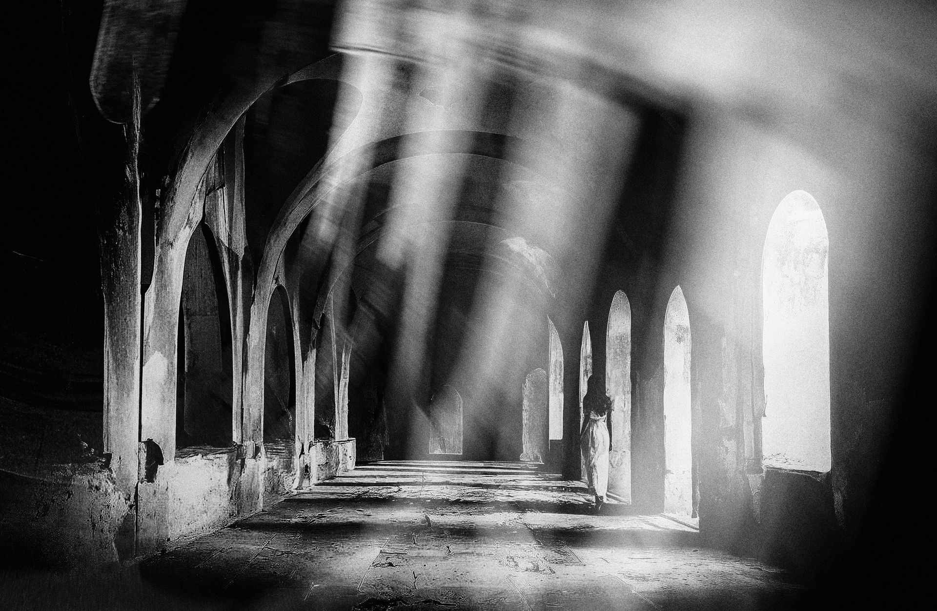 Foto Art in schwarz weiß. Eine Frau mit weißem Kleid geht durch ein lichtdurchflutetes altes zerfallenes Gewölbe