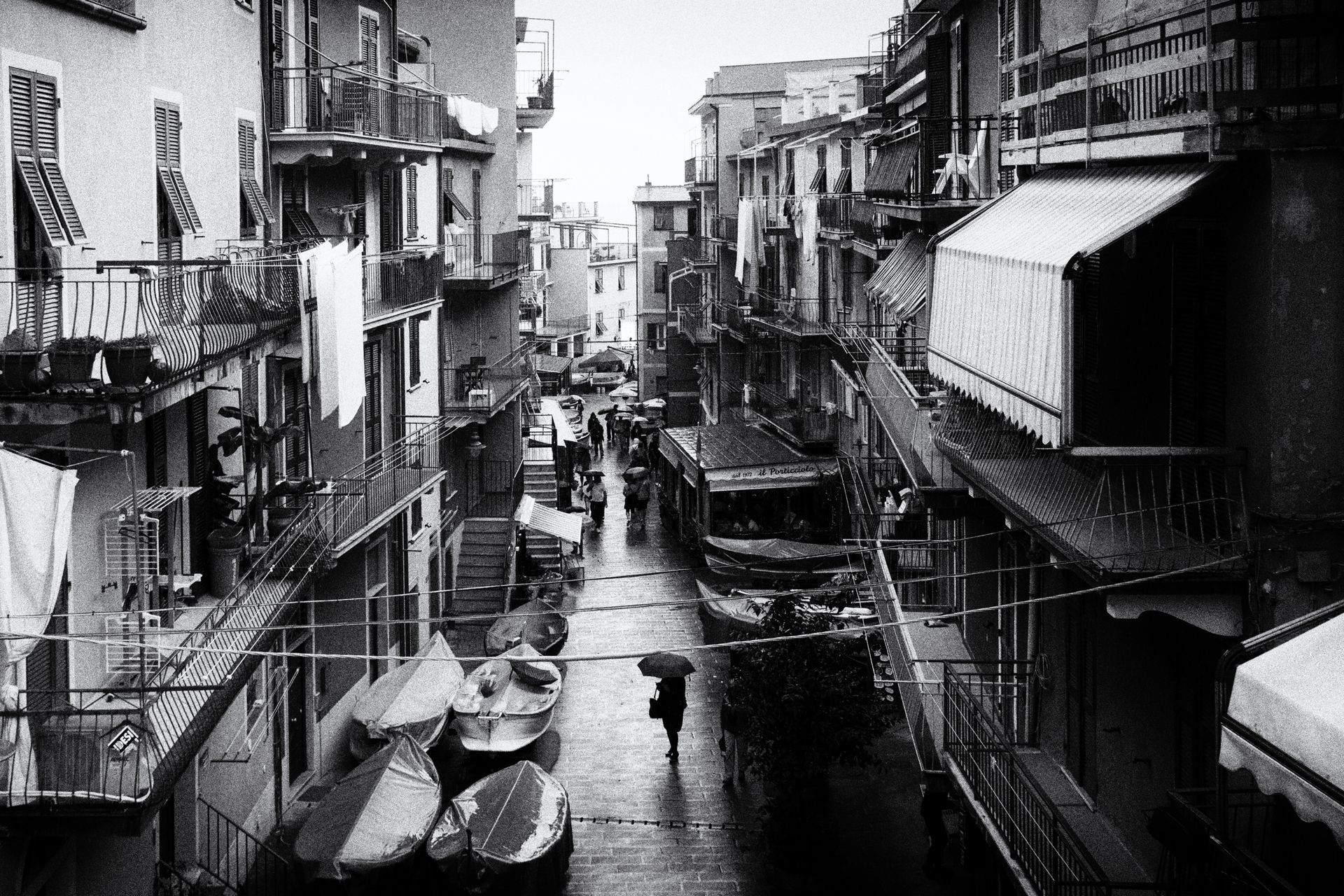 Eine Frau mit Regenschirm steht in der Mitte einer Straße in Manarola Cinque Terre und schaut auf abgestellte kleine Fischerboote. Straßenfotografie aus Italien in schwarz weiss.