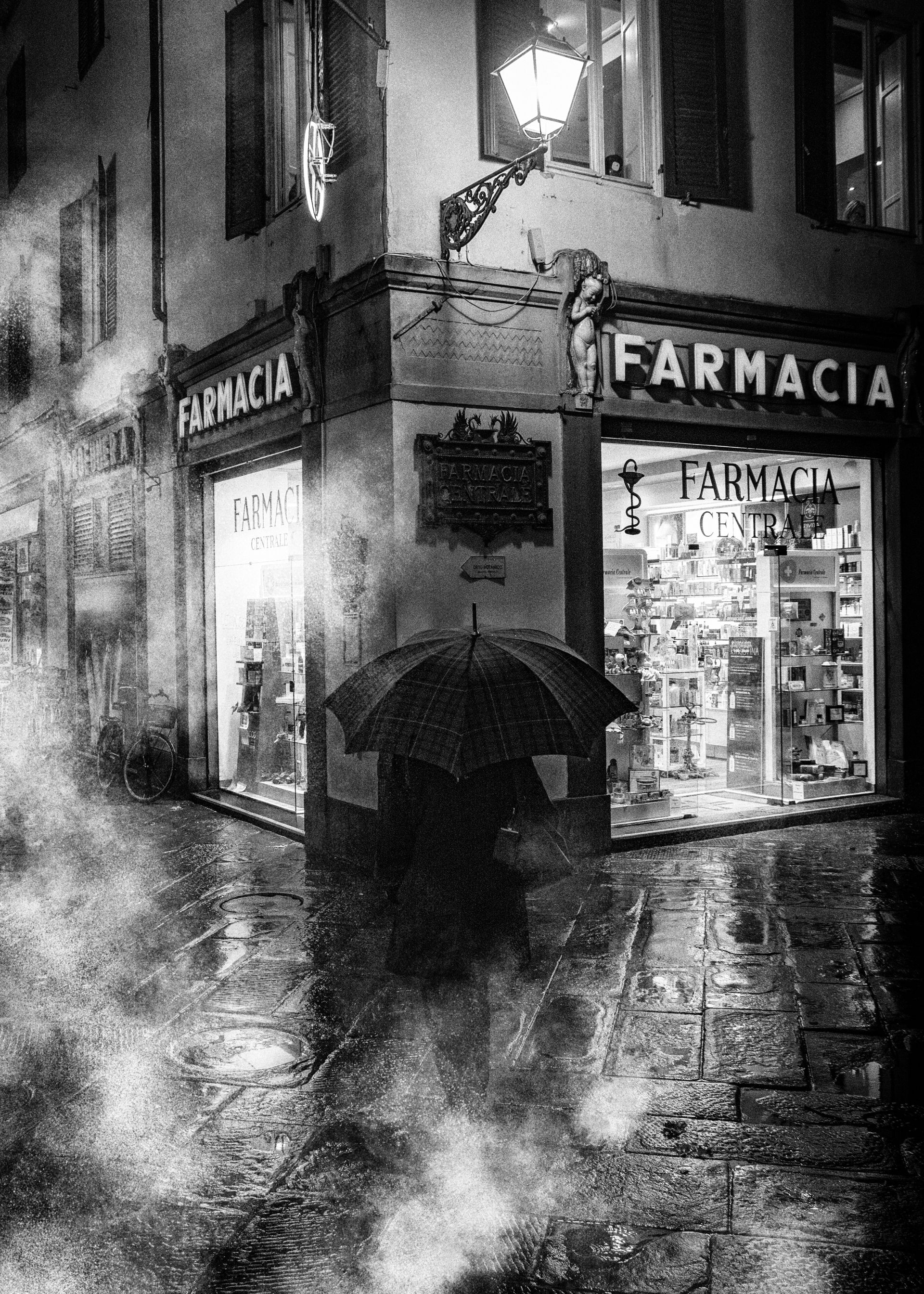 Eine Frau mit Regenschirm geht allein durch die Altstadt von Lucca in einer regnerischen Nacht. Straßenfotografie aus Italien in schwarz weiss.