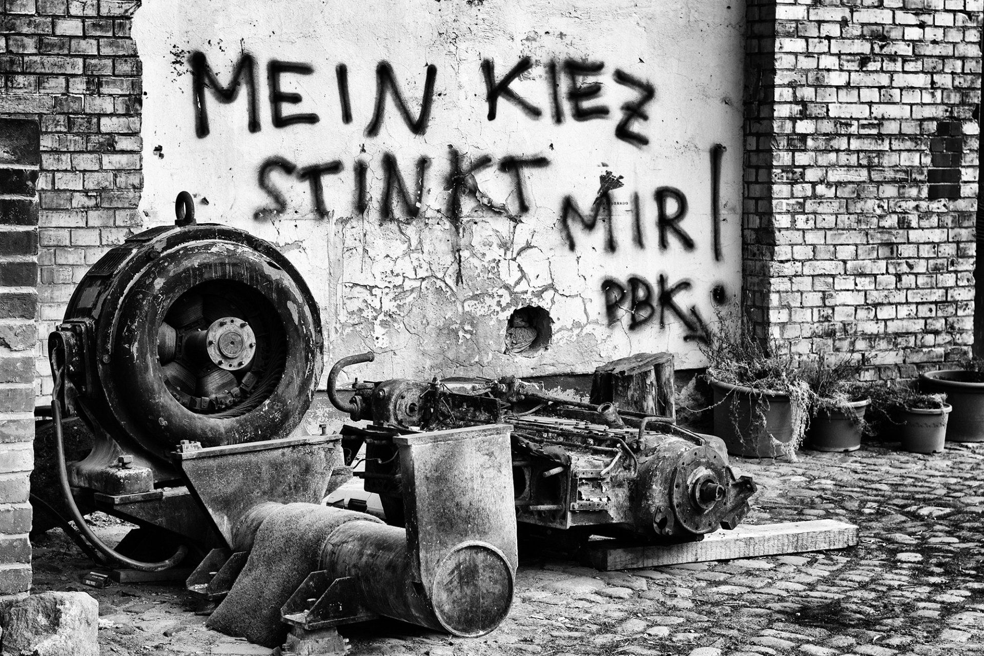 Graffiti an einer alten Hauswand hinter verrosteten Maschinenteilen in Berlin Prenzlauer Berg. Straßenfotografie aus Berlin schwarz und weiß.