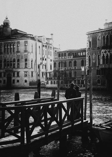Ein junges Paar steht auf einem Bootssteg am Canal Grande und schaut in die Altstadt von Venedig. Street Photography aus Italien in schwarz weiß.
