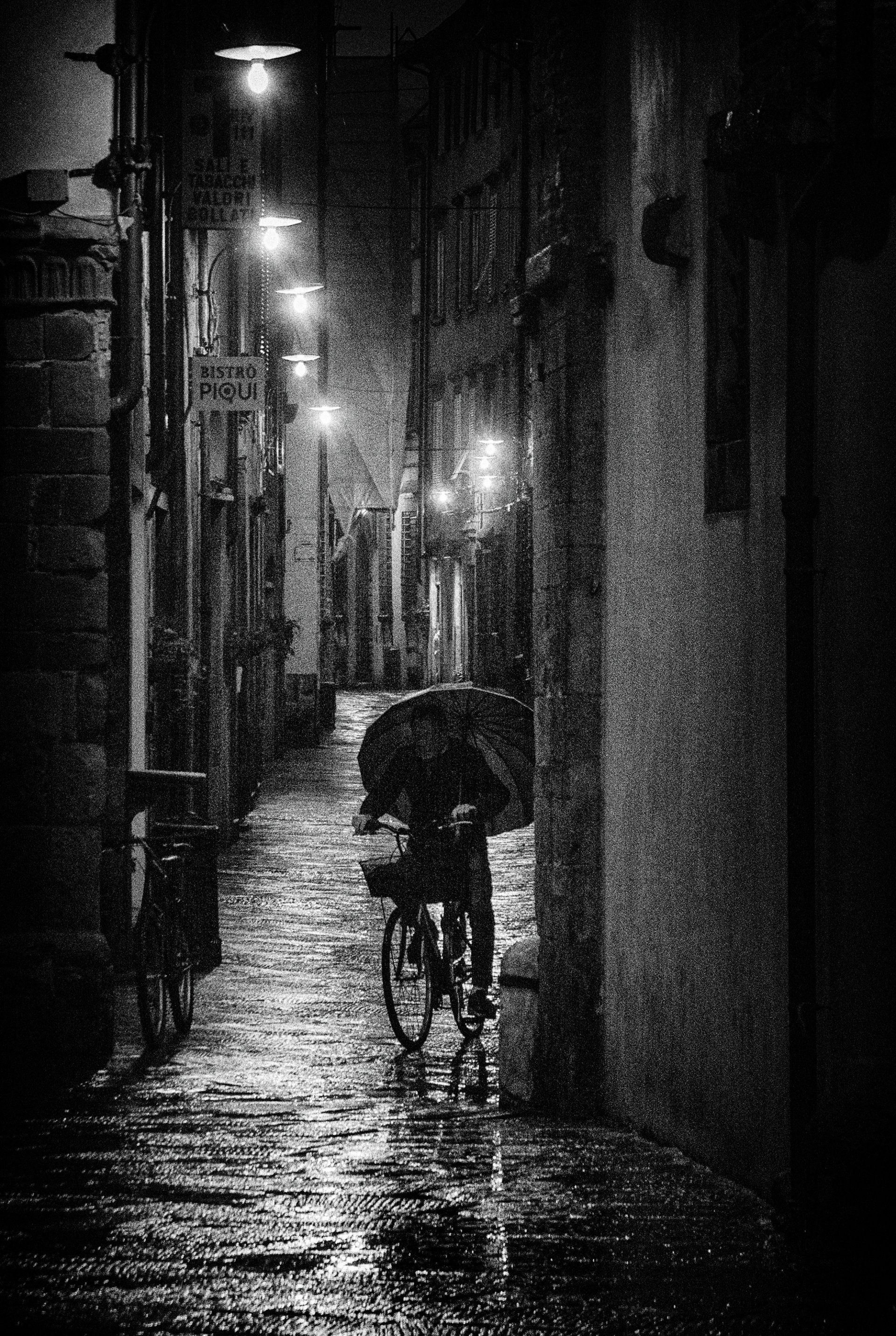 Ein Fahrradfahrer mit Regenschirm fährt Nachts durch eine beleuchtete Straße in der Altstadt von Lucca. Straßenfotografie aus Italien in schwarz weiß.