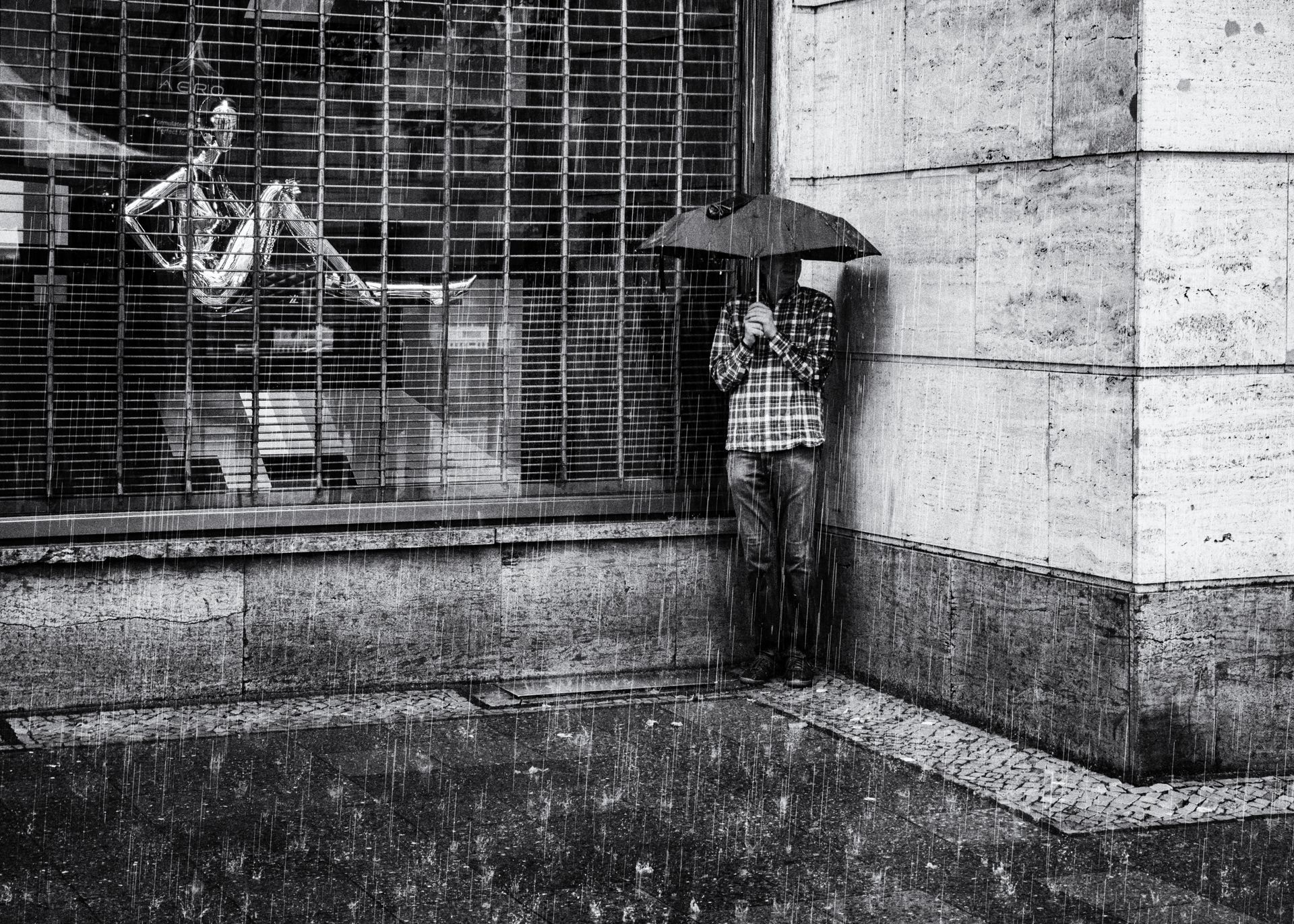 Ein Mann mit Regenschirm steht in einer Hausecke und versucht sich vor heftigem Regen zu schützen. Straßenfotografie aus Berlin in schwarz weiß.