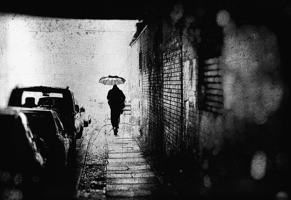 An einem regenerischen Tag in Berlin geht ein Mann mit Regenschirm eilig durch einen kleinen Tunnel. Abstrakte Straßenfotografie aus Berlin in schwarz weiß.