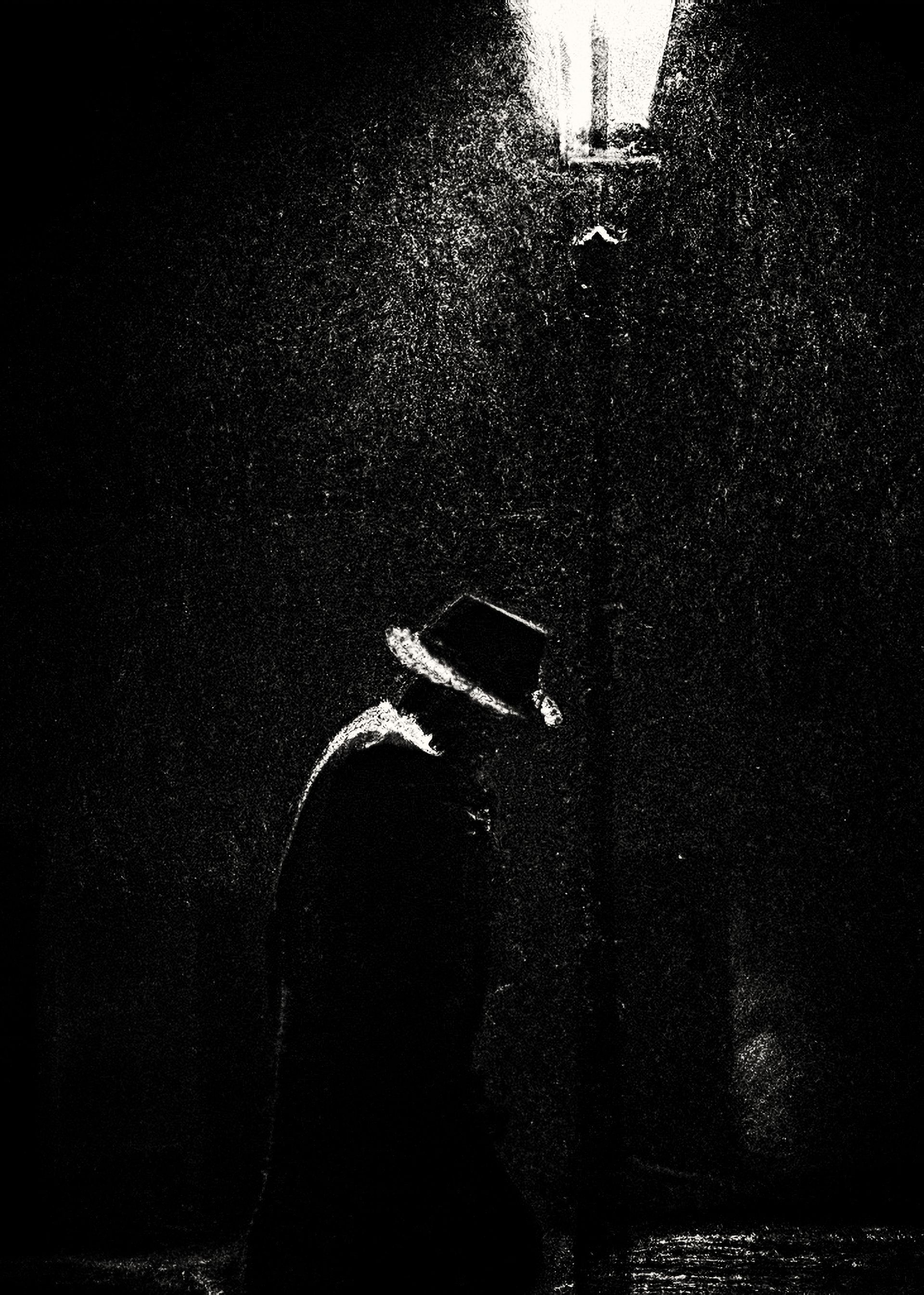 Schwarz weiß Fotografie eines Mannes mit Hut und Mantel nachts unter einer leuchtenden Laterne bei starken Regen