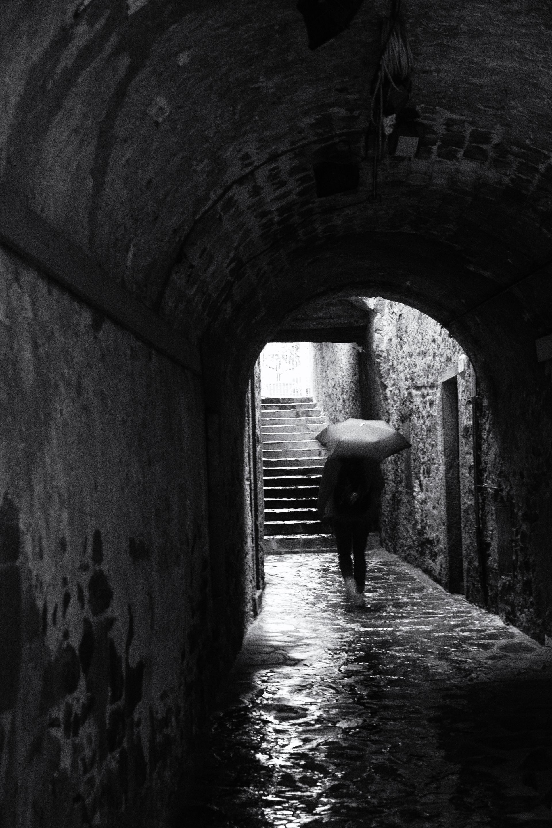 Schwarz weiß Fotografie einer Frau mit Regenschirm, die an einem regnerischen Tag in der Toskana eilig durch einen regennassen Hausdurchgang geht